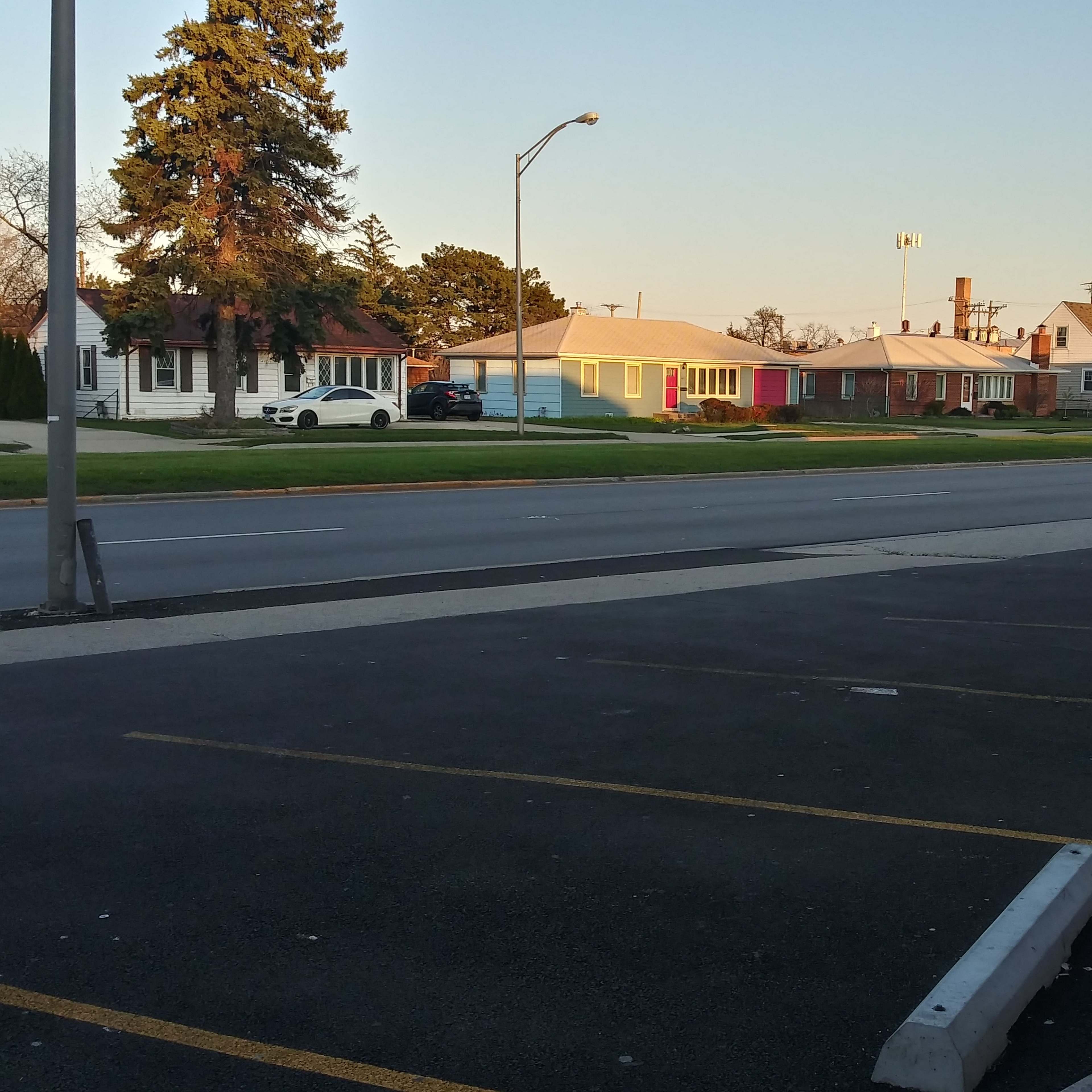 A quiet street is lined with various houses on one side and a parking lot on the other, with a few cars parked along the curb.