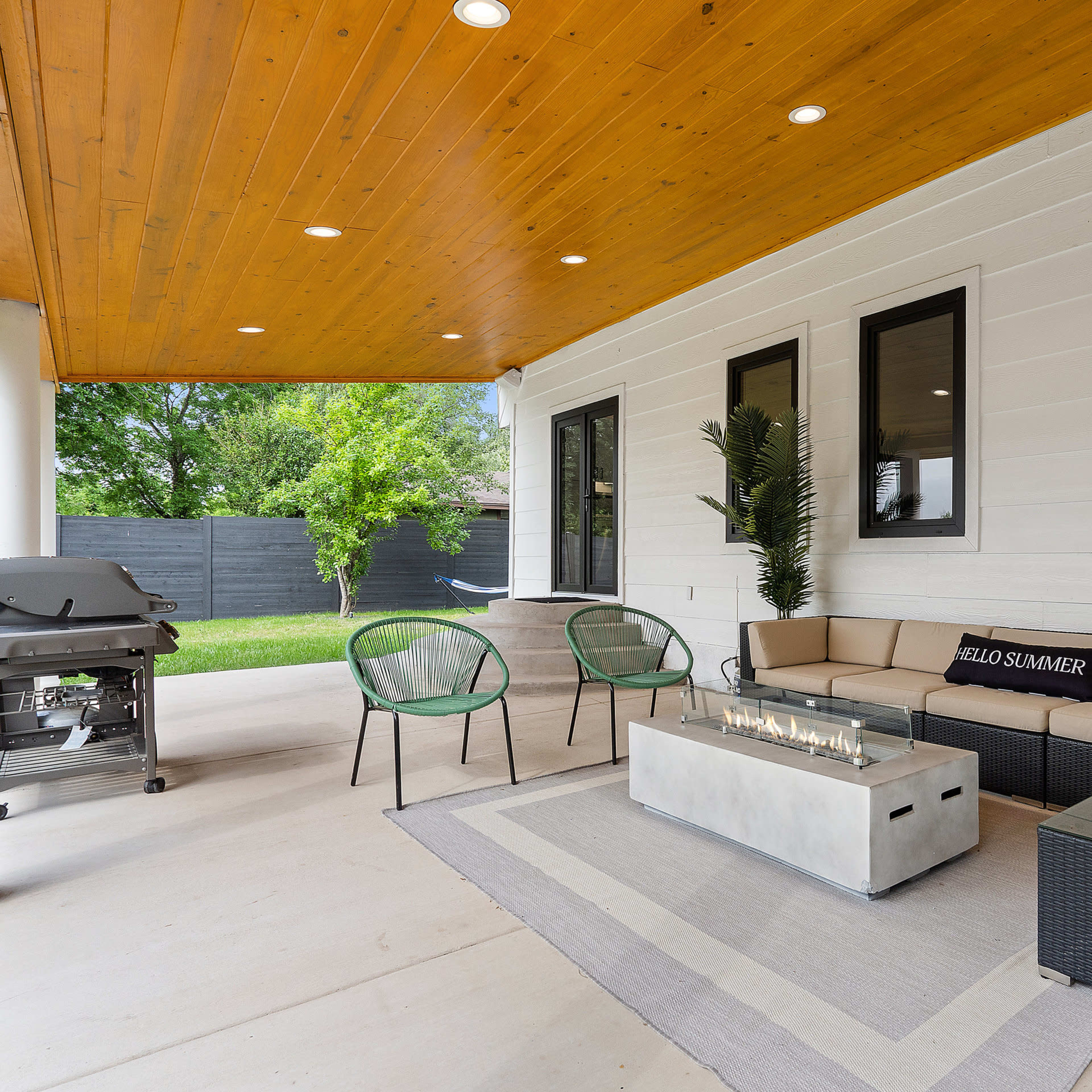 A covered outdoor seating area features a sectional sofa, a coffee table with a fire feature, and green chairs under a wooden ceiling.