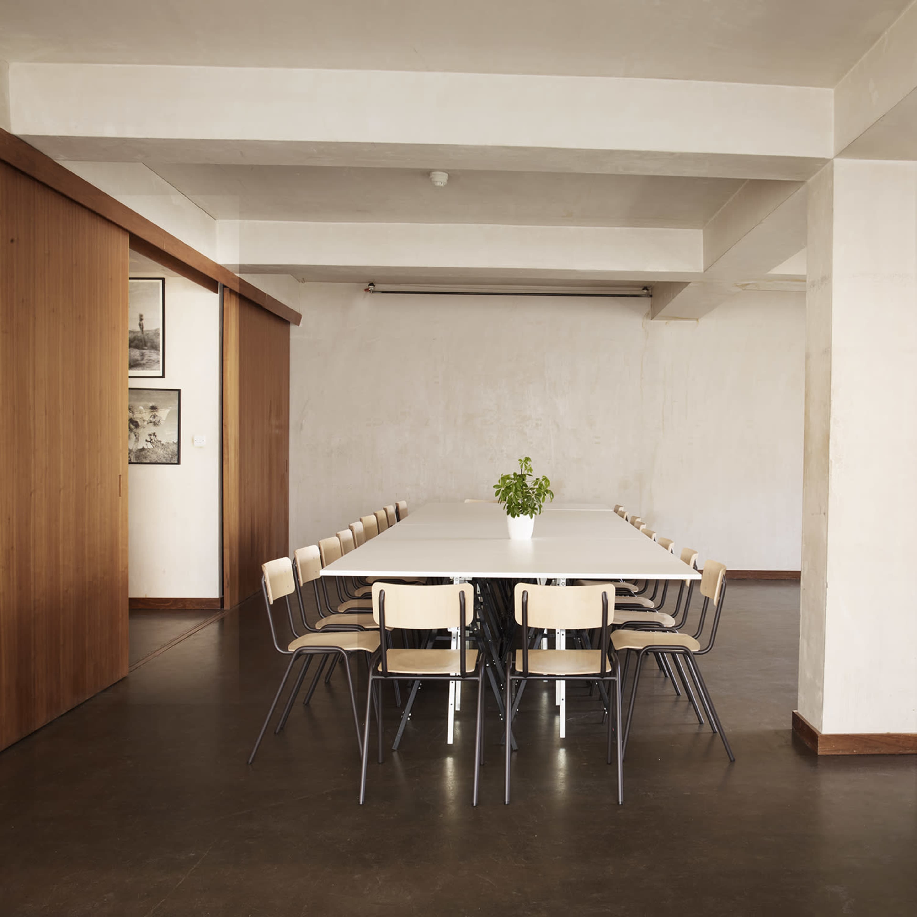 A large, empty conference table surrounded by wooden chairs is set in a spacious, minimalist room with plants and wooden accents.