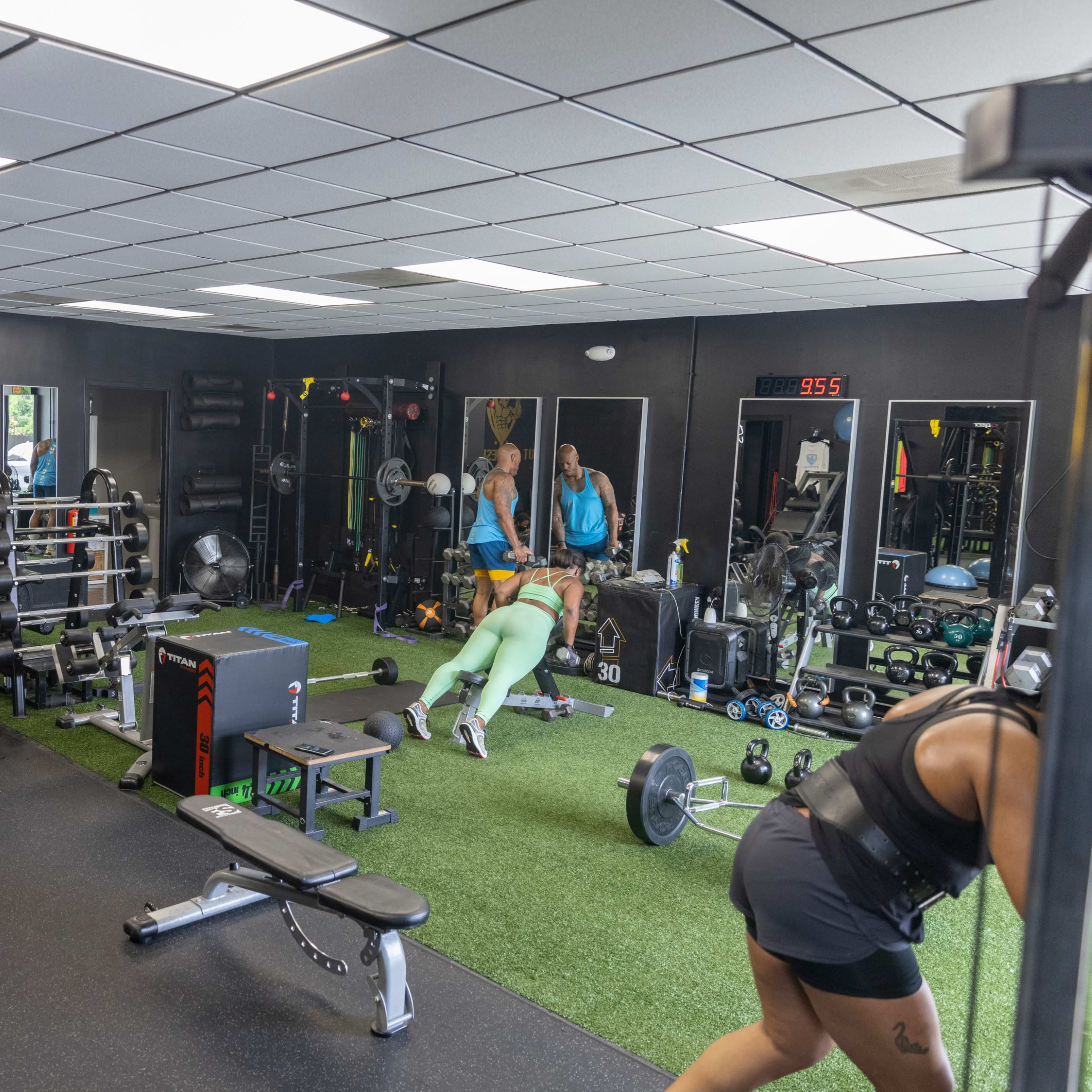 A gym interior with various exercise equipment, including weights and benches, and two individuals engaged in workouts on a green turf area.