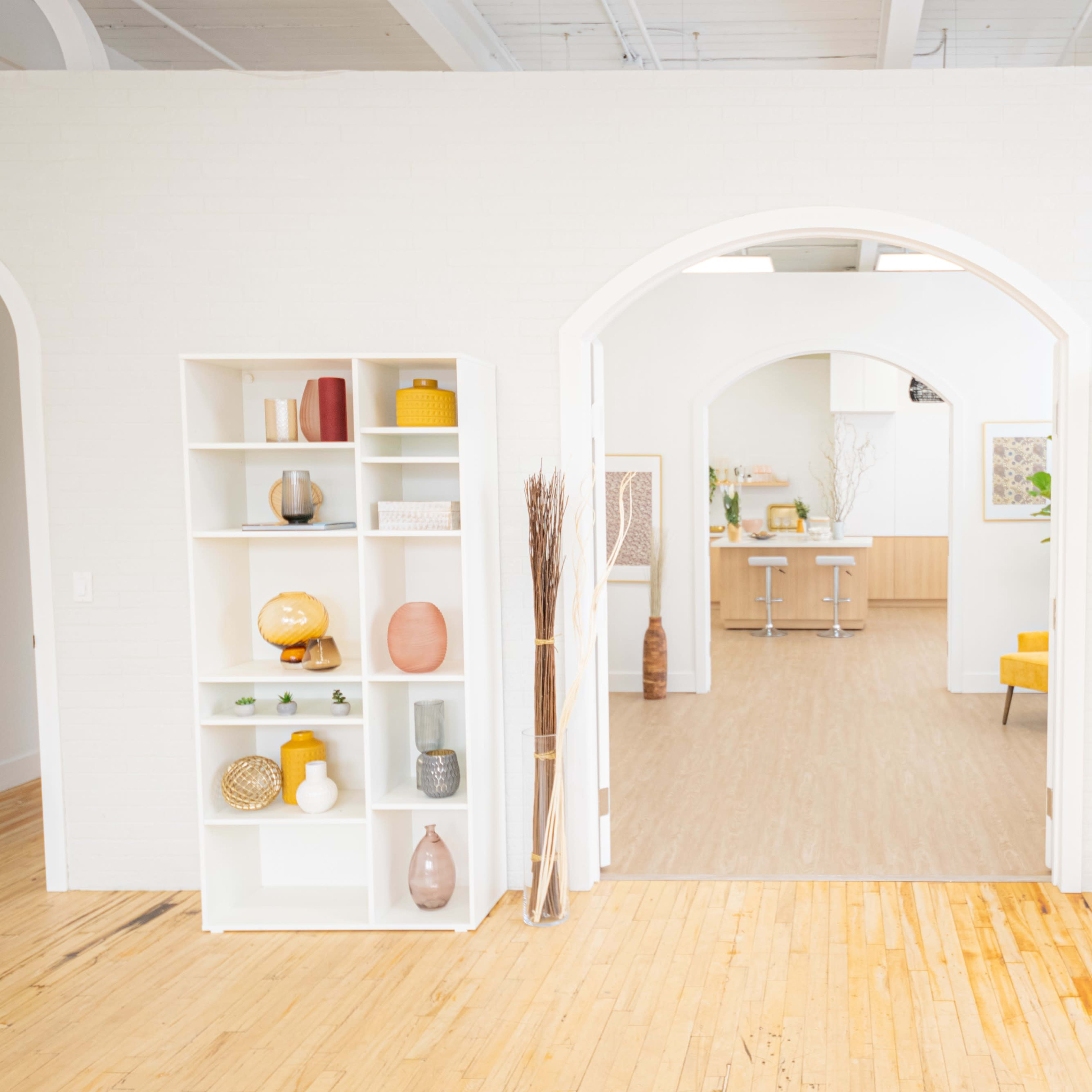 A bright room with two white bookshelves on either side of an open archway, leading to a light-furnished area with wooden flooring and minimalistic decor.