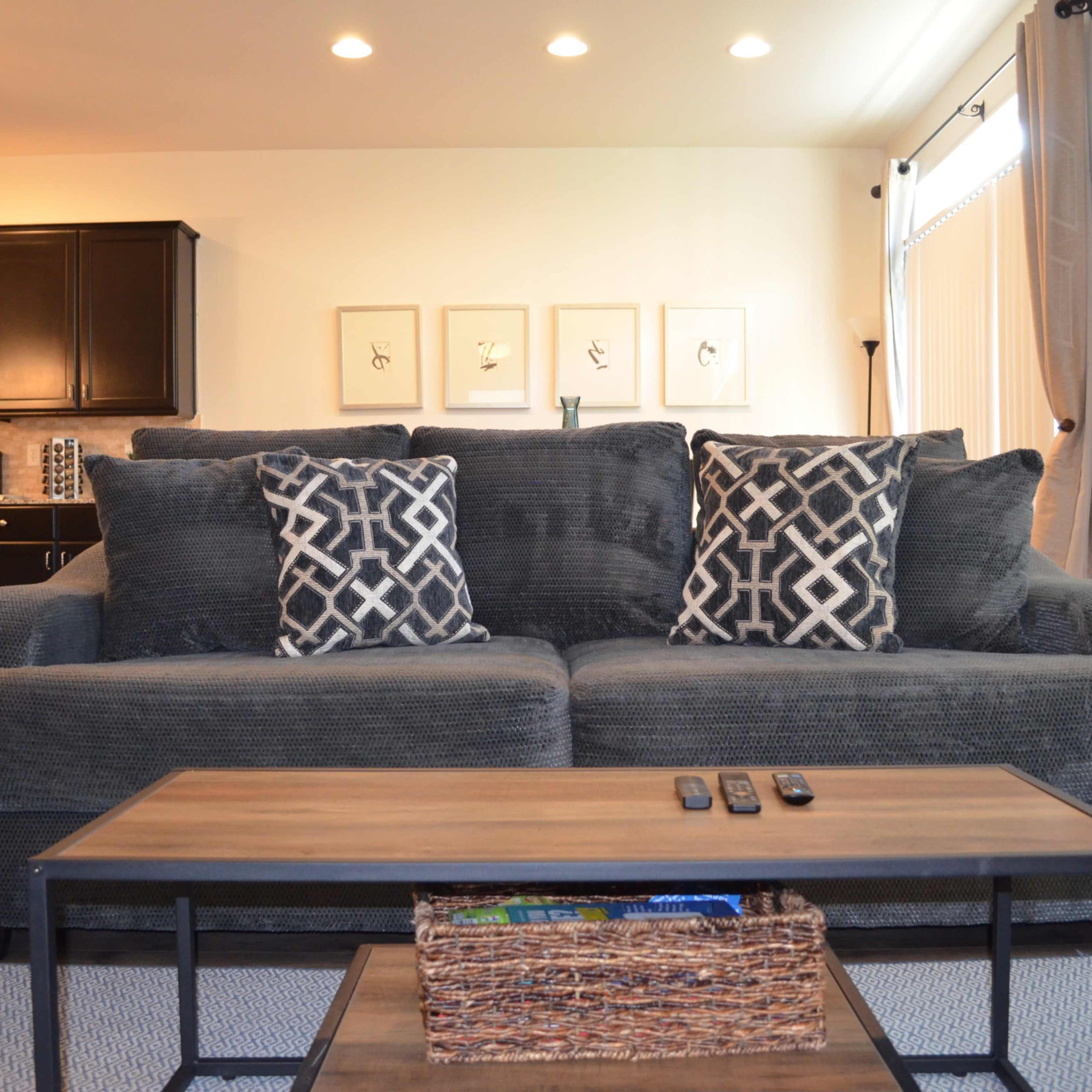 A grey sofa with patterned cushions is positioned in a living room featuring a coffee table with a basket and remote controls in front of it.