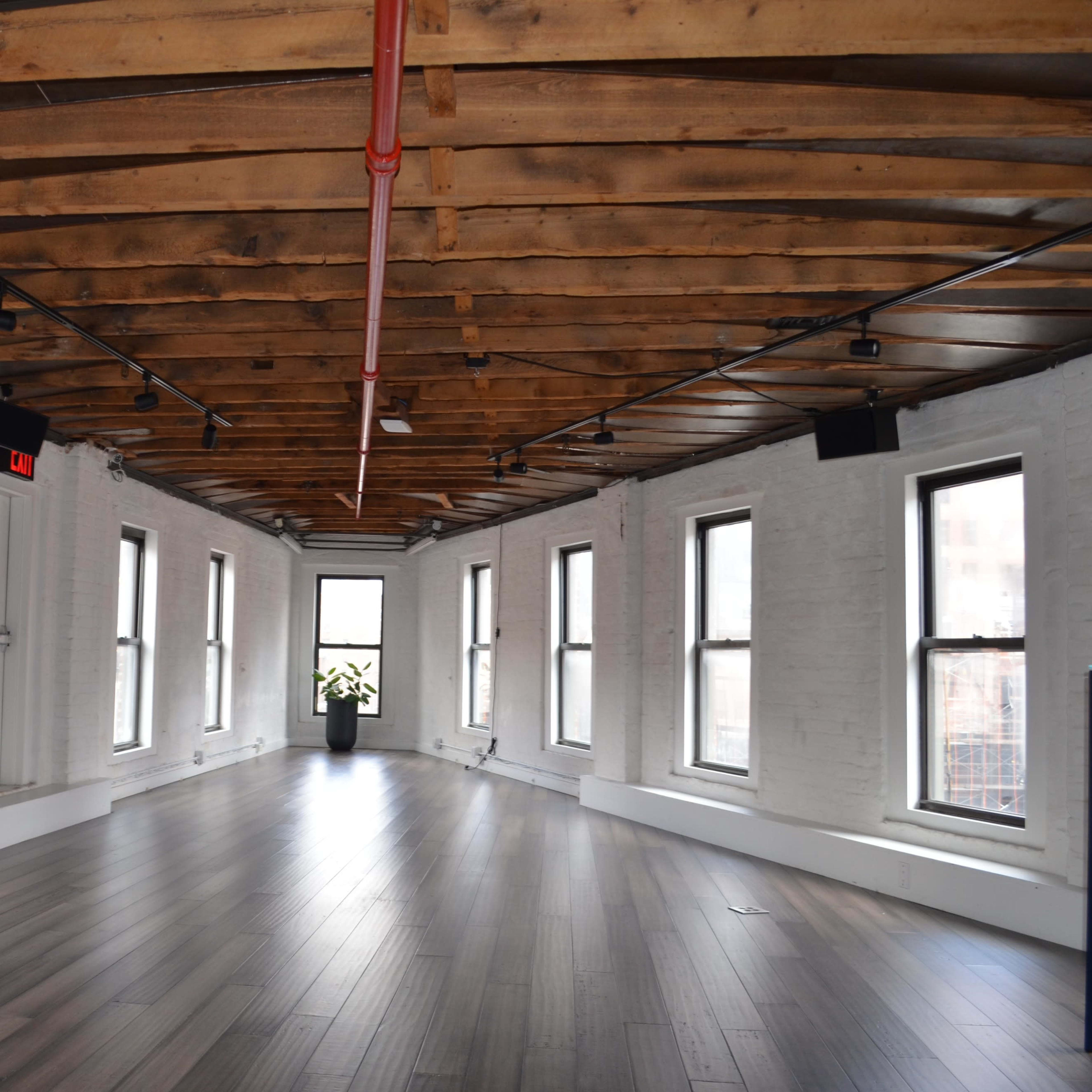 A spacious room with wooden beams and large windows, featuring a bench along one wall and a potted plant in the corner.