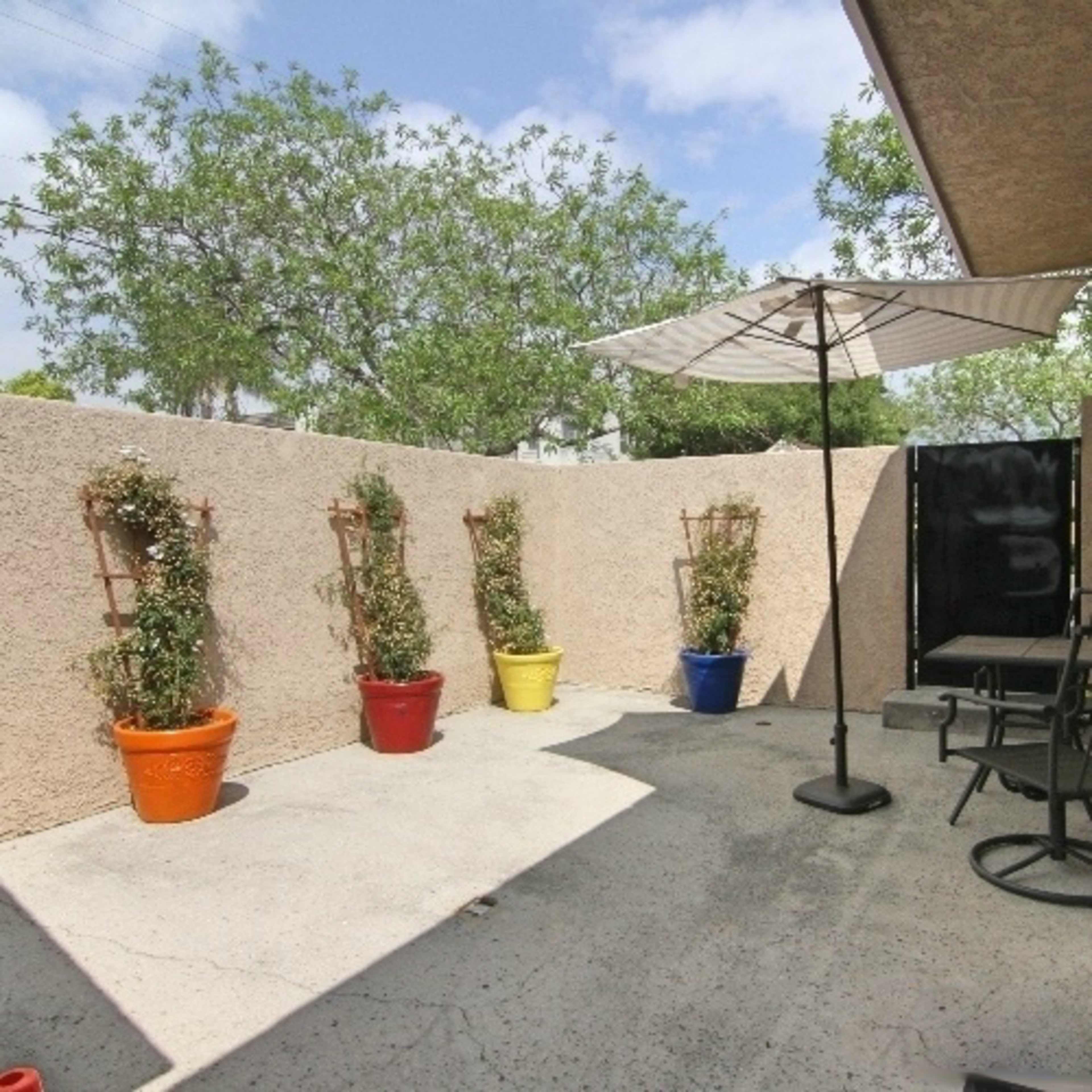 A small, enclosed outdoor patio features potted plants in colorful containers, a table with a parasol, and a couple of black chairs against a textured wall.
