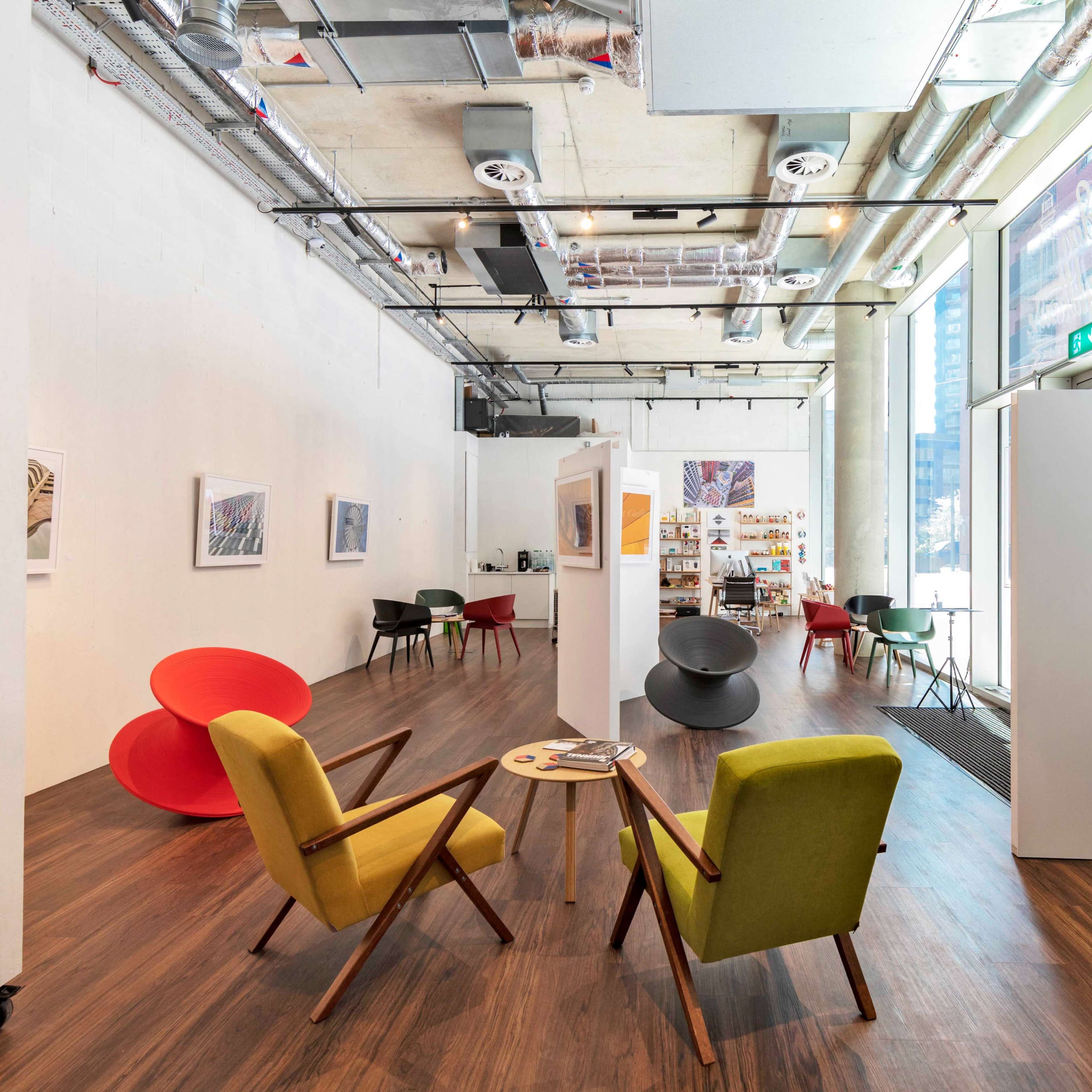 An indoor space with wooden flooring, featuring modern furniture arrangements, including chairs and a small table, along with white partition walls and artwork displayed on the walls.