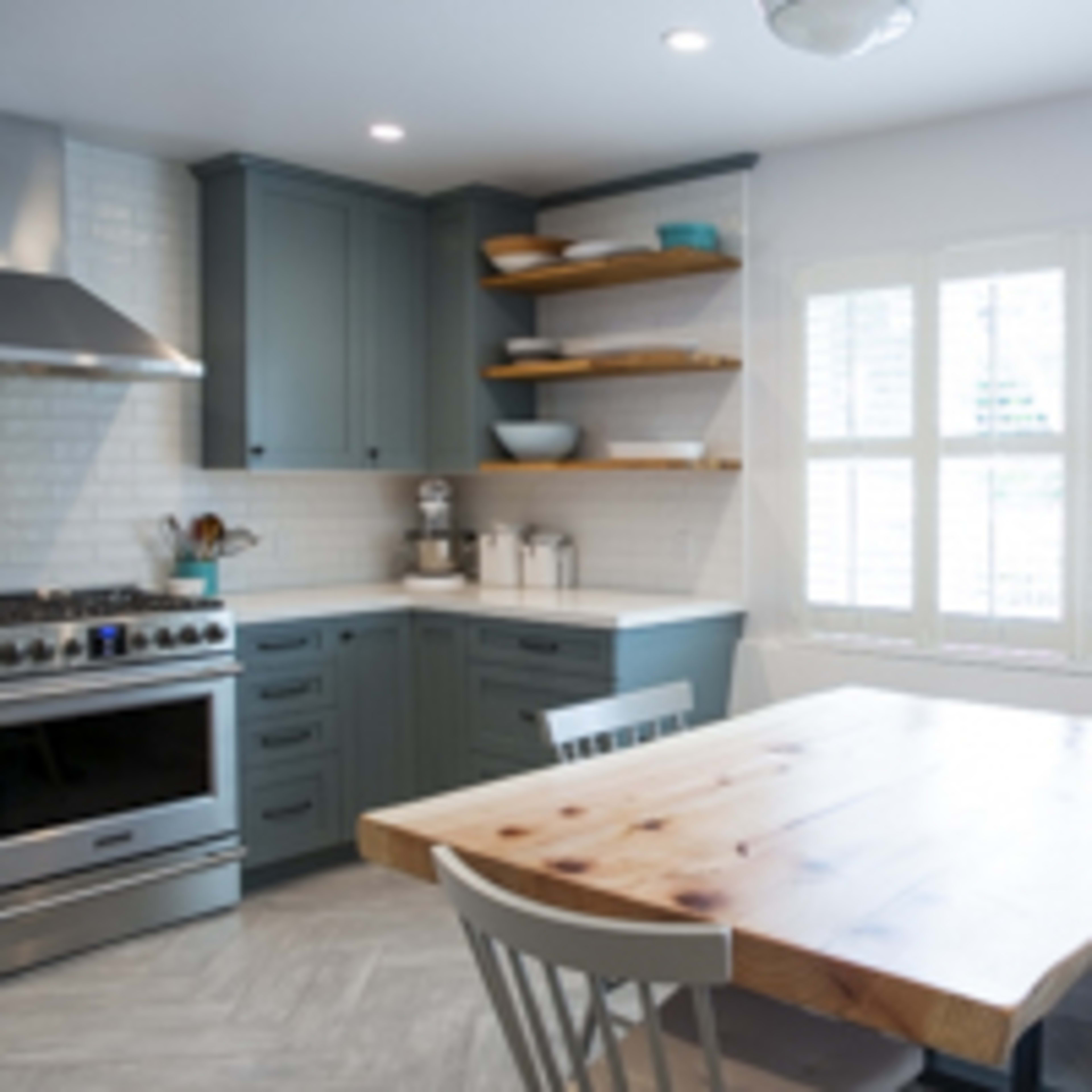 A modern kitchen features a stainless steel stove, blue cabinetry, open shelves with dishware, and a wooden dining table with grey seating next to a window.