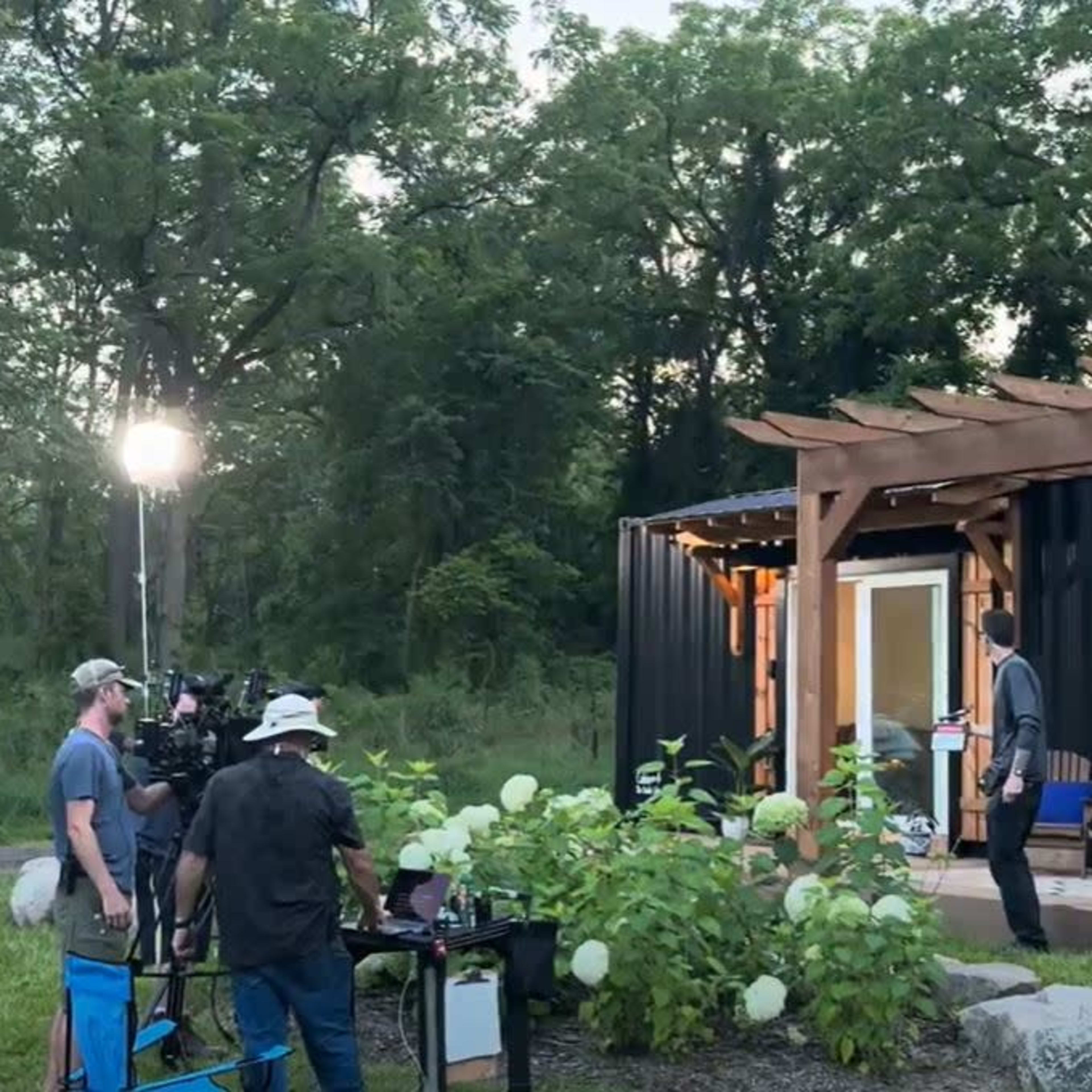 A film crew sets up cameras and equipment outside a black container-style building amidst greenery.