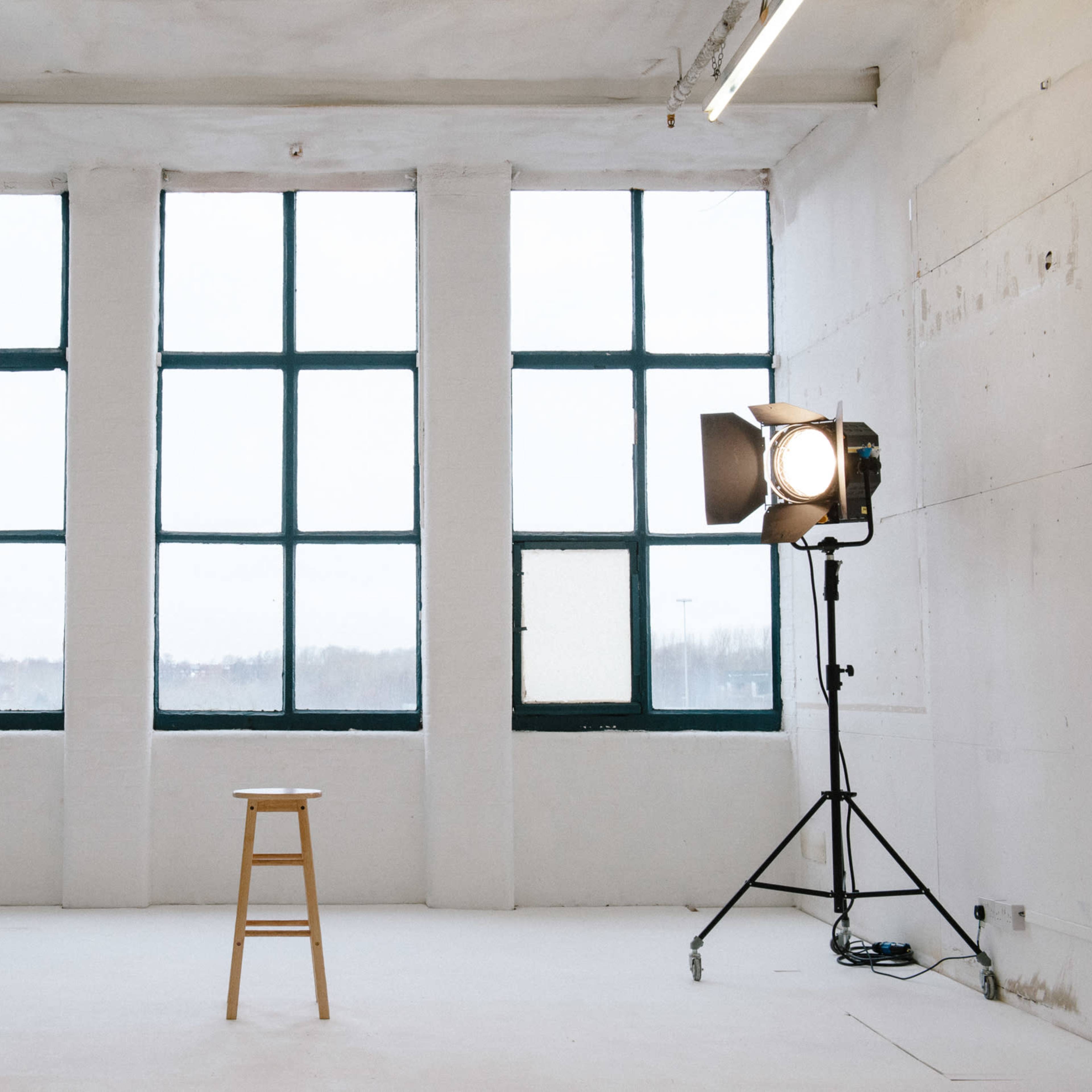 A minimalist studio space with large windows, a wooden stool in the center, and a light setup on a stand beside a stepladder.