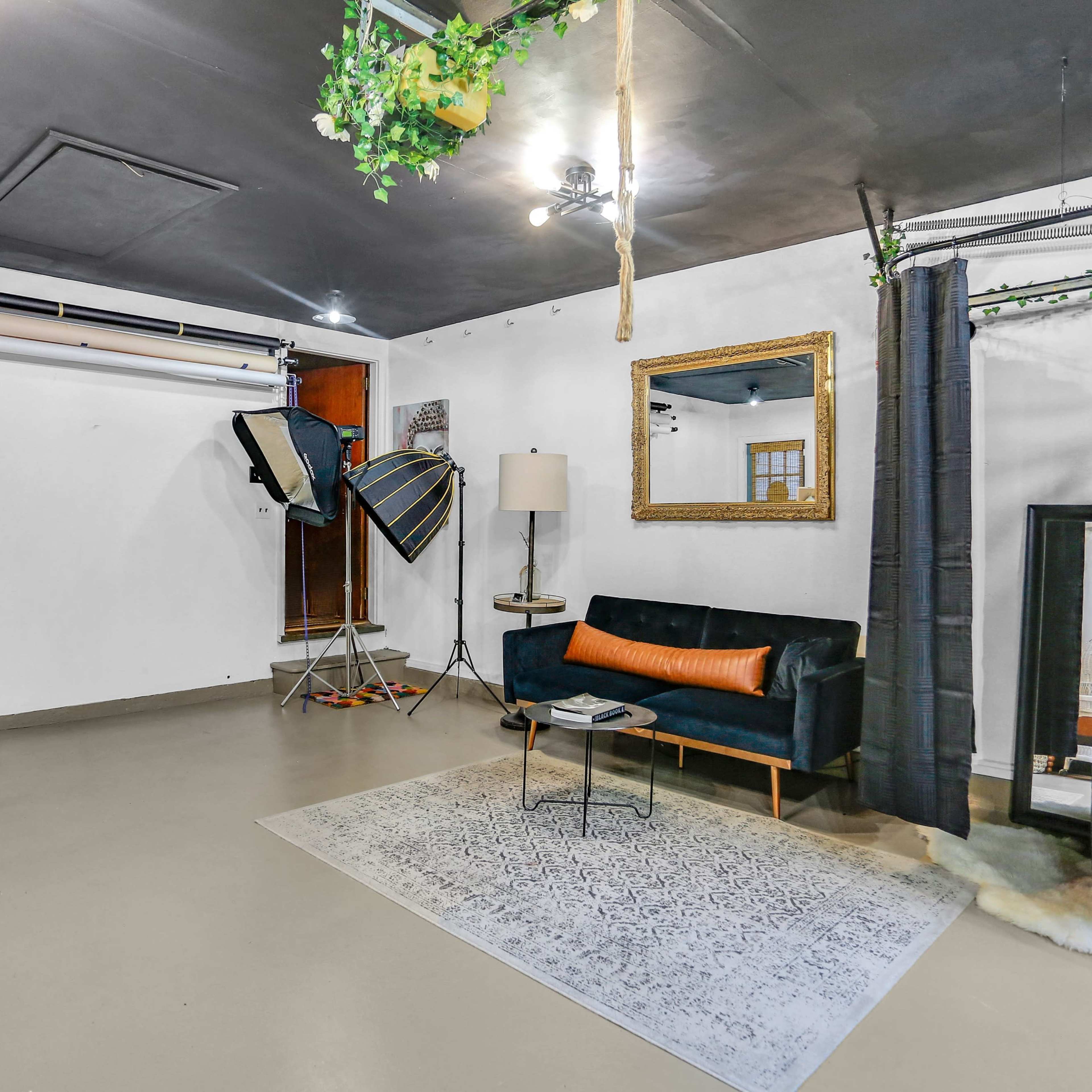 A spacious room with a black sofa, a standing lamp, a large mirror, a patterned rug, and photography equipment including softboxes and backdrops.