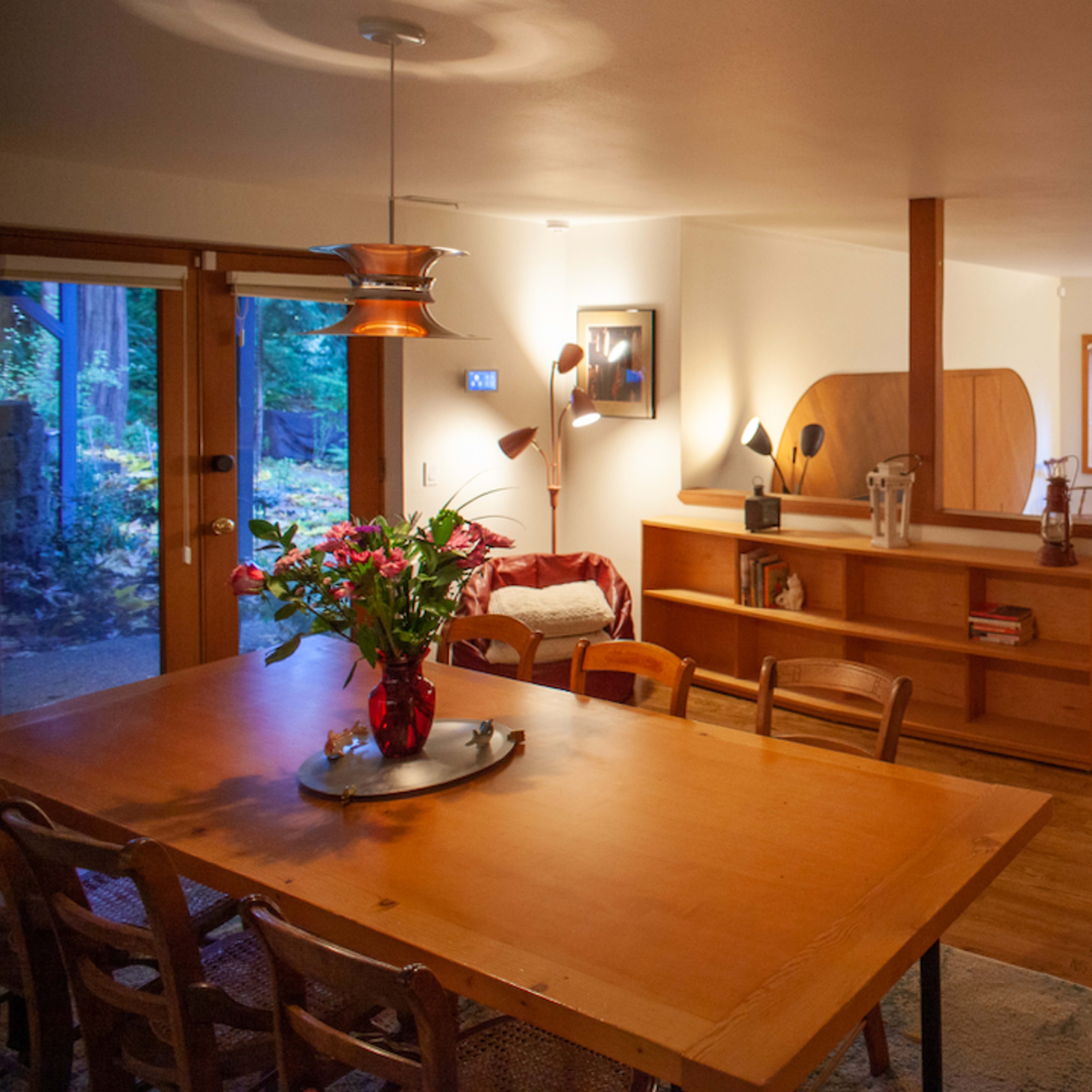 A wooden dining table is set in a well-lit open space, leading into a cozy living area with large windows overlooking a wooded view.