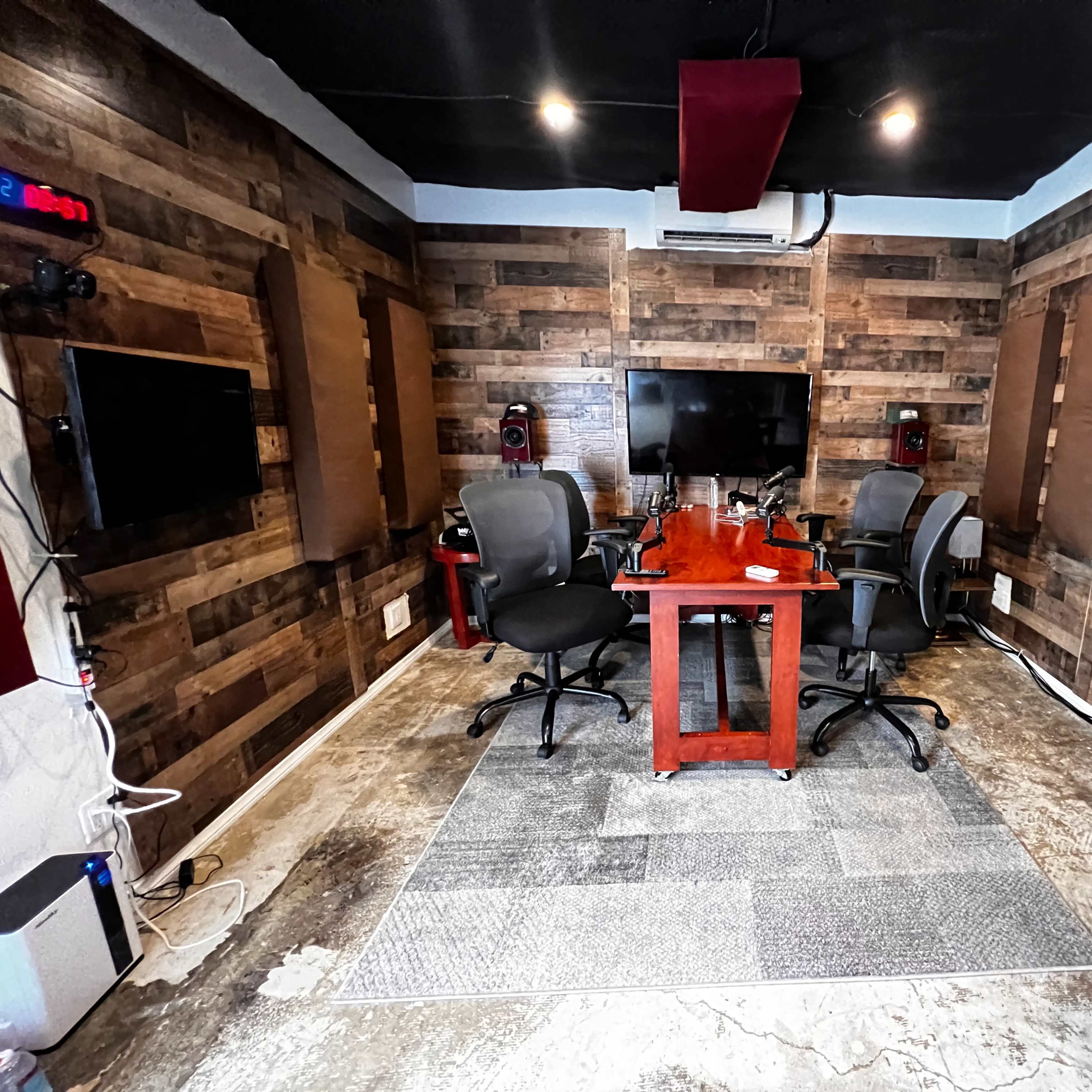 A small conference room features a wooden table surrounded by four chairs, with two wall-mounted screens and a large monitor against a wood-paneled backdrop.