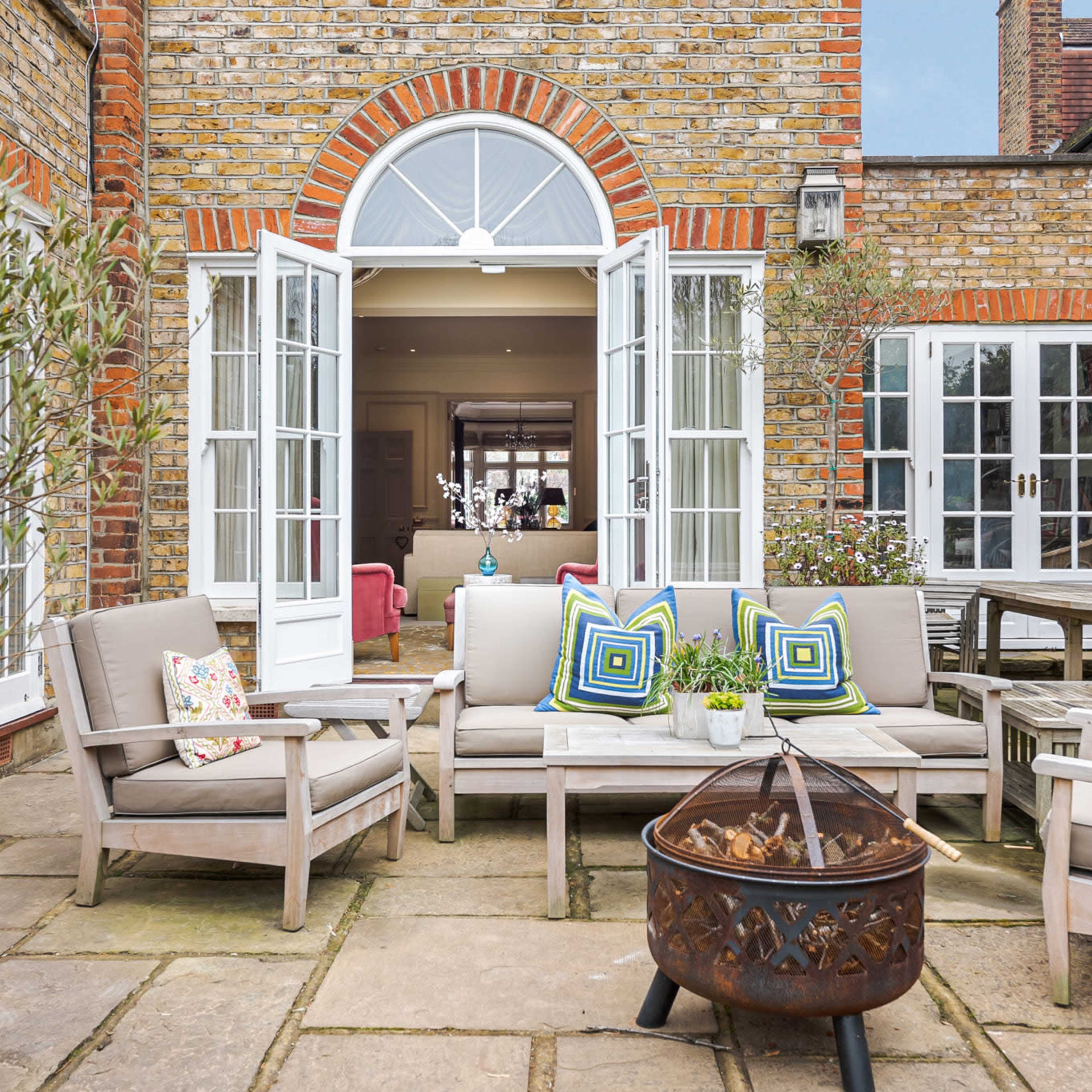 An outdoor patio area with wooden furniture, colorful cushions, and a fire pit, surrounded by brick walls and open double doors leading into a room.