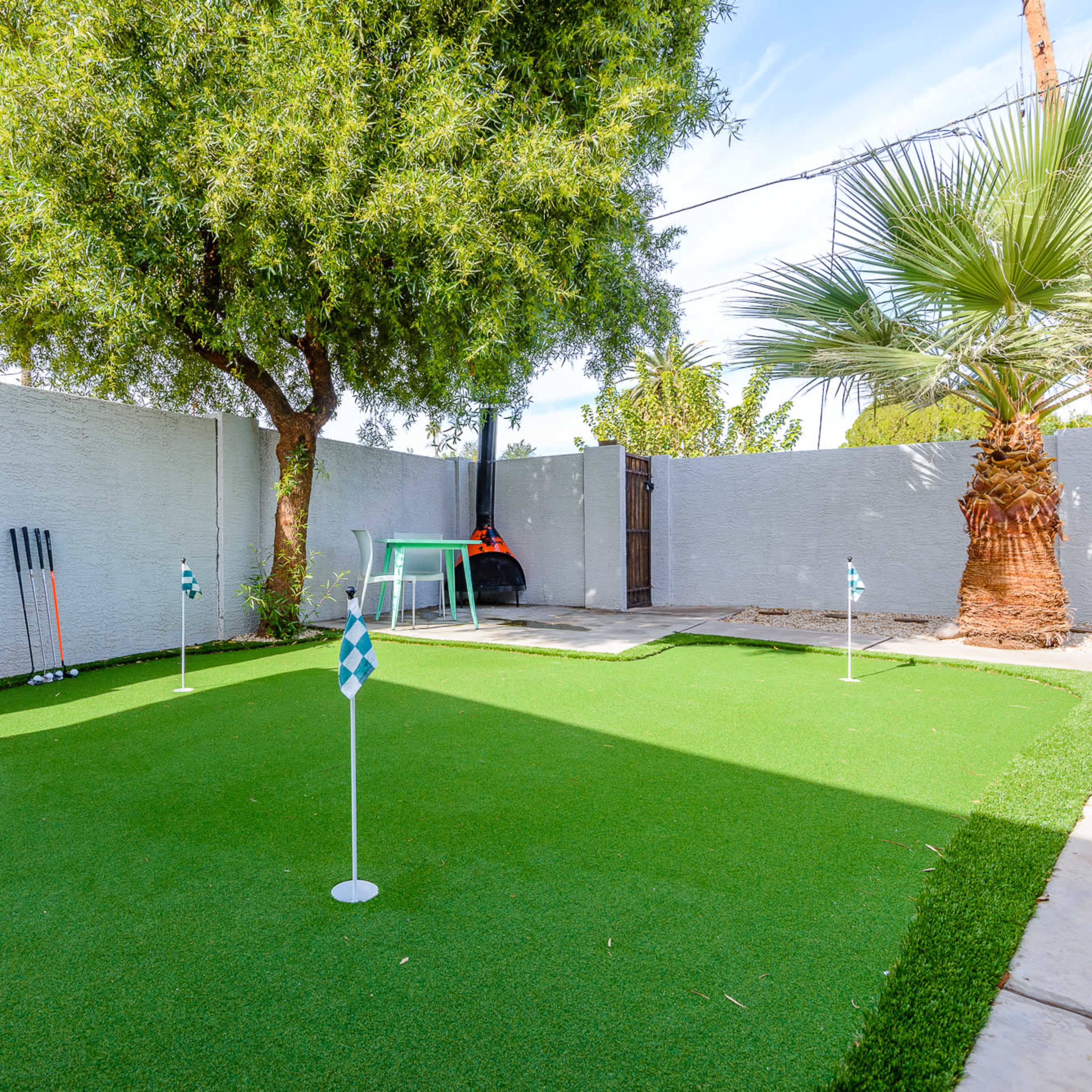 A small backyard putting green surrounded by a tree, palm, and a patio area.