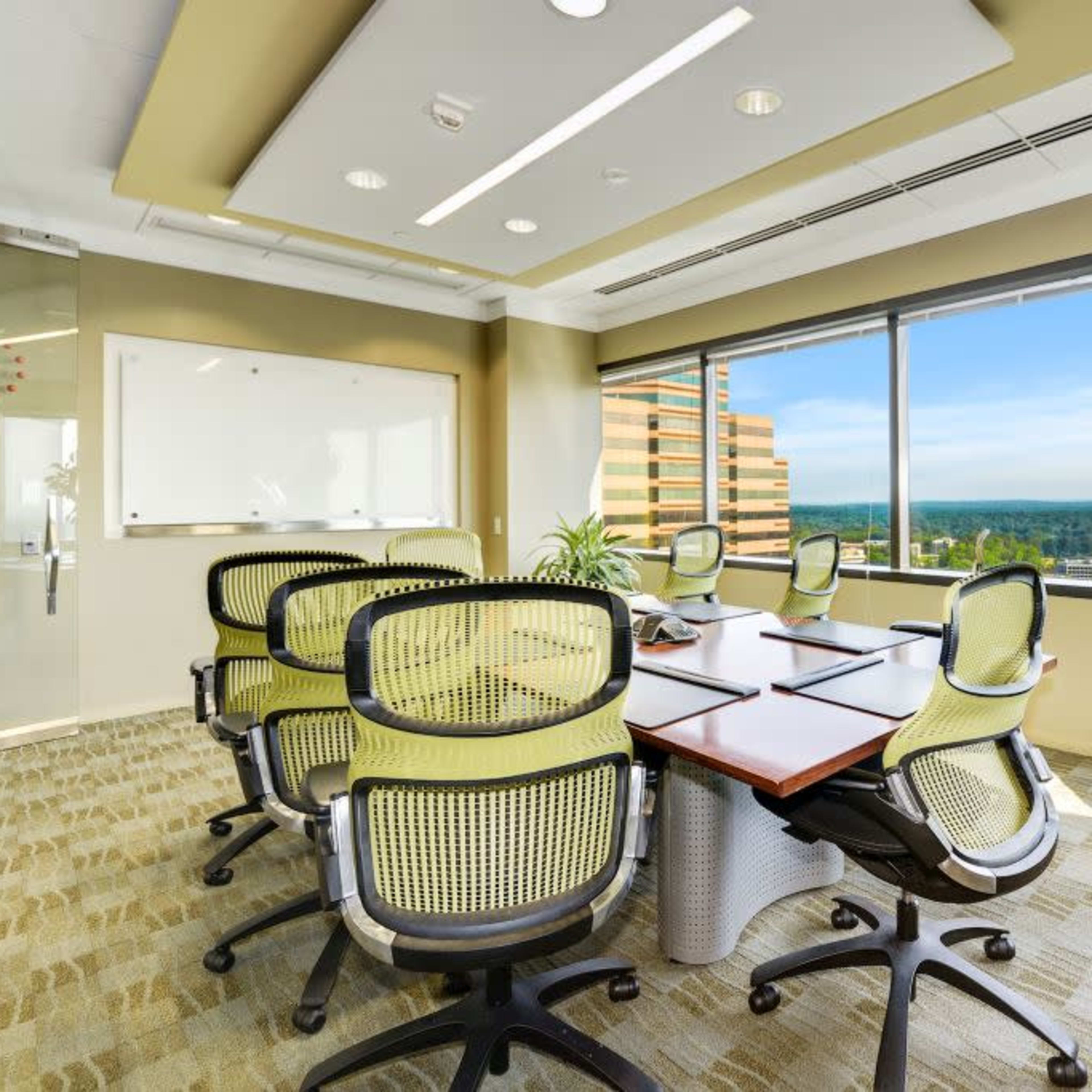 A modern conference room features a large, oval table surrounded by six green office chairs and large windows offering a view of the landscape outside.