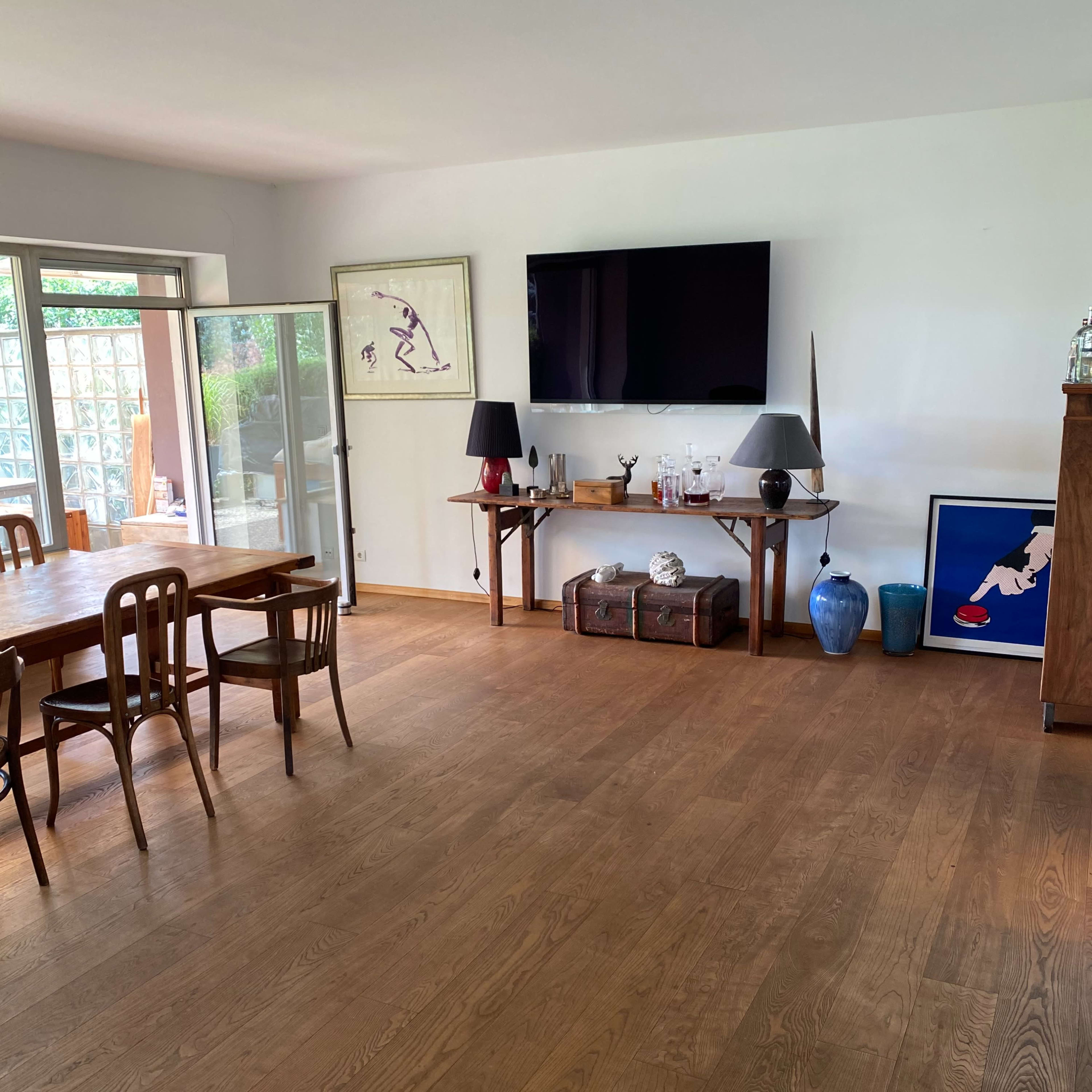 A modern living room with wooden flooring, a large television mounted on the wall, a dining table with chairs, and various decorative items on display.