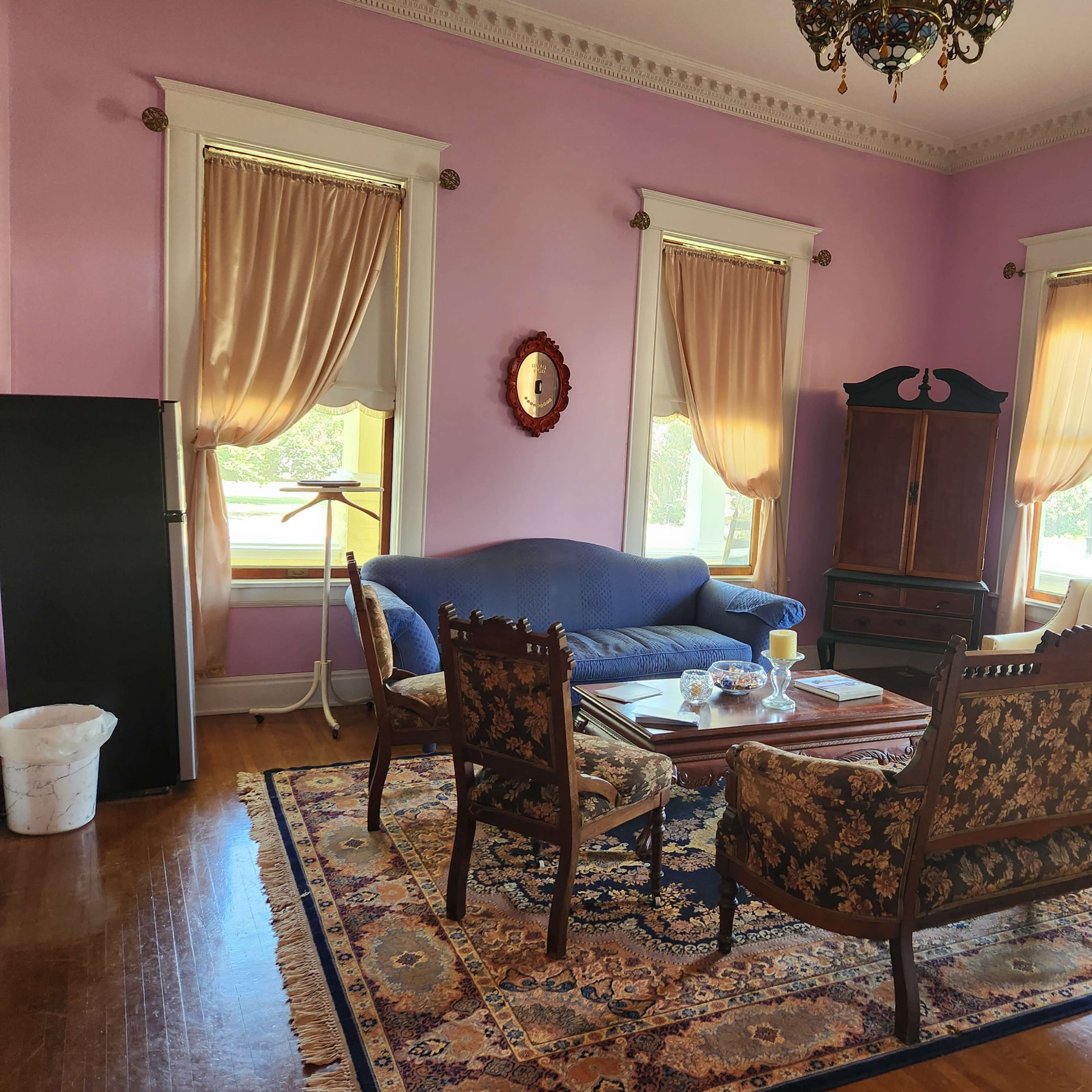 The room features a pink wall color, a blue couch, a cream-colored sofa, and dark wooden furniture arranged around a central coffee table on a large area rug.