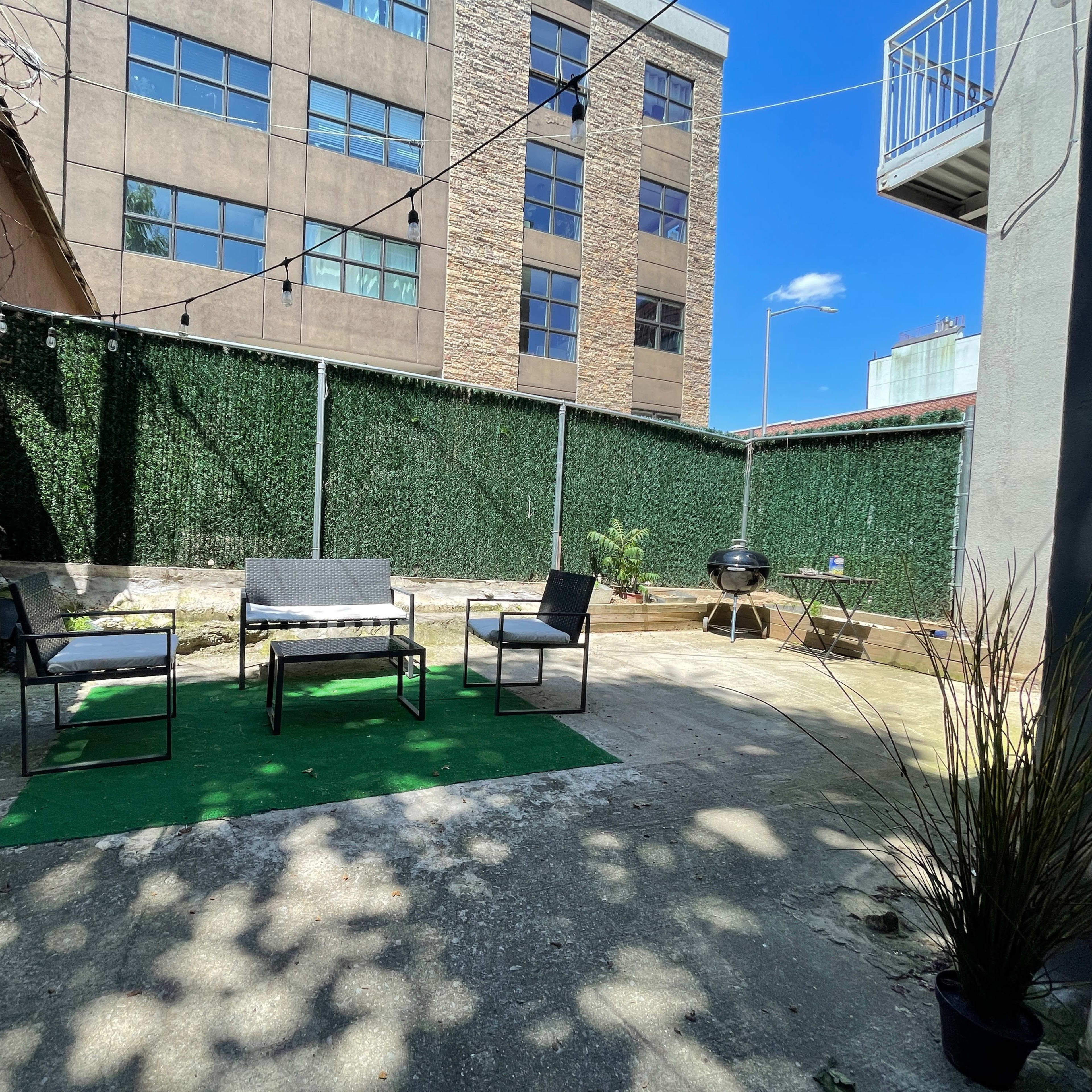 A small outdoor patio area with a green rug, four chairs arranged around a coffee table, a grill, and a backdrop of a wall covered in greenery.