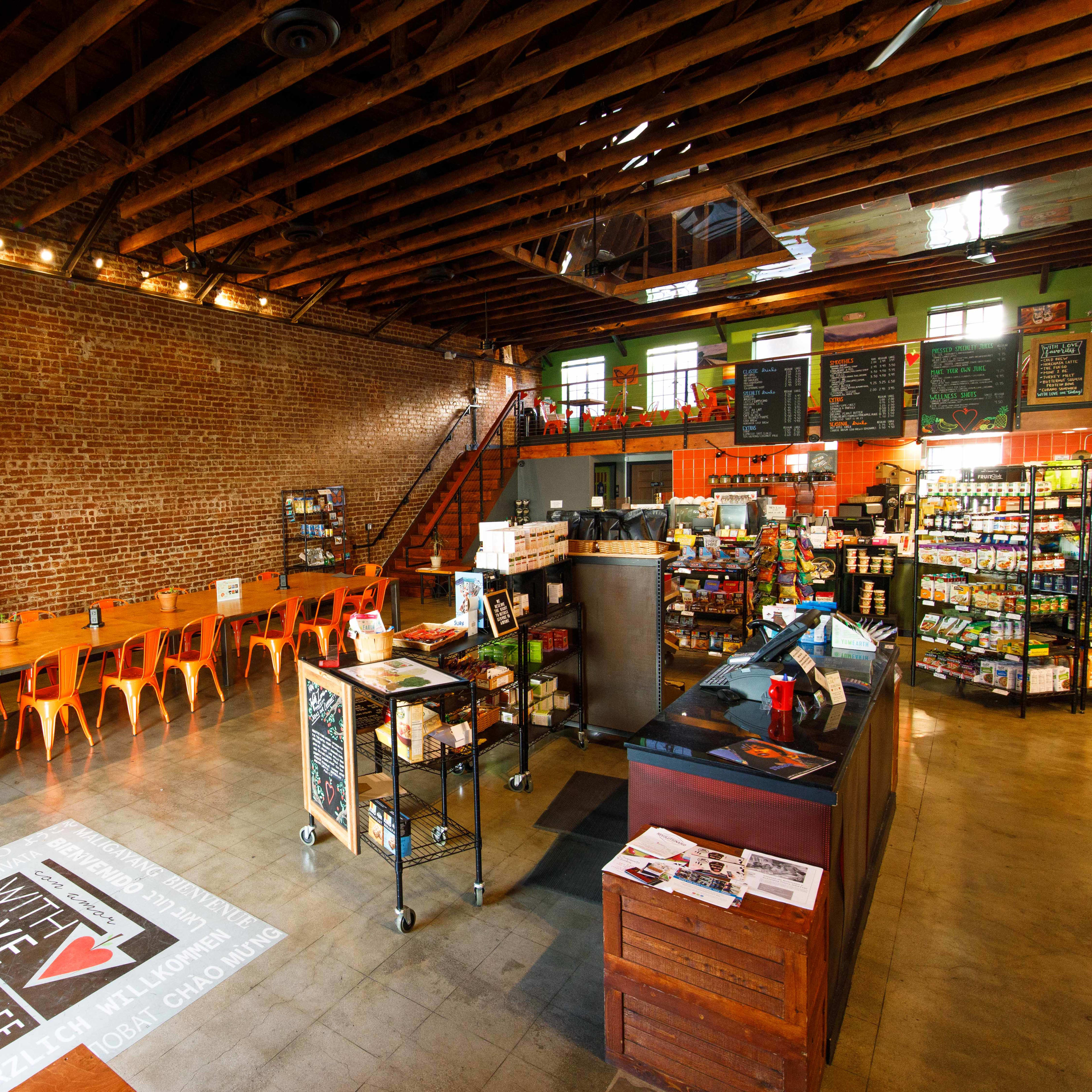 A spacious café features wooden tables and orange chairs along a brick wall, with shelves of products and a service counter in the foreground.