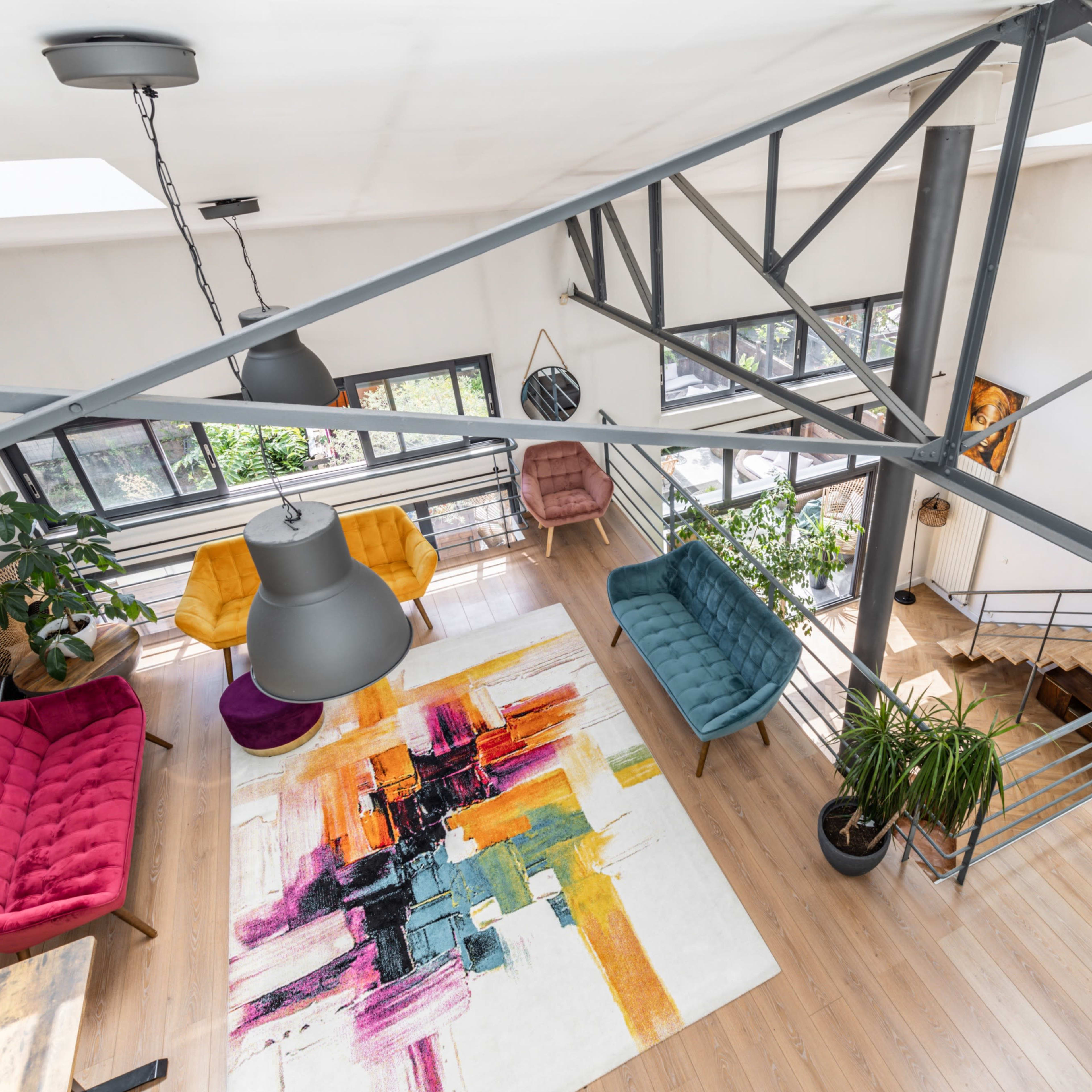 A spacious living area with colorful furniture, a large abstract rug, and a staircase leading to a lower level, all under a skylit ceiling.