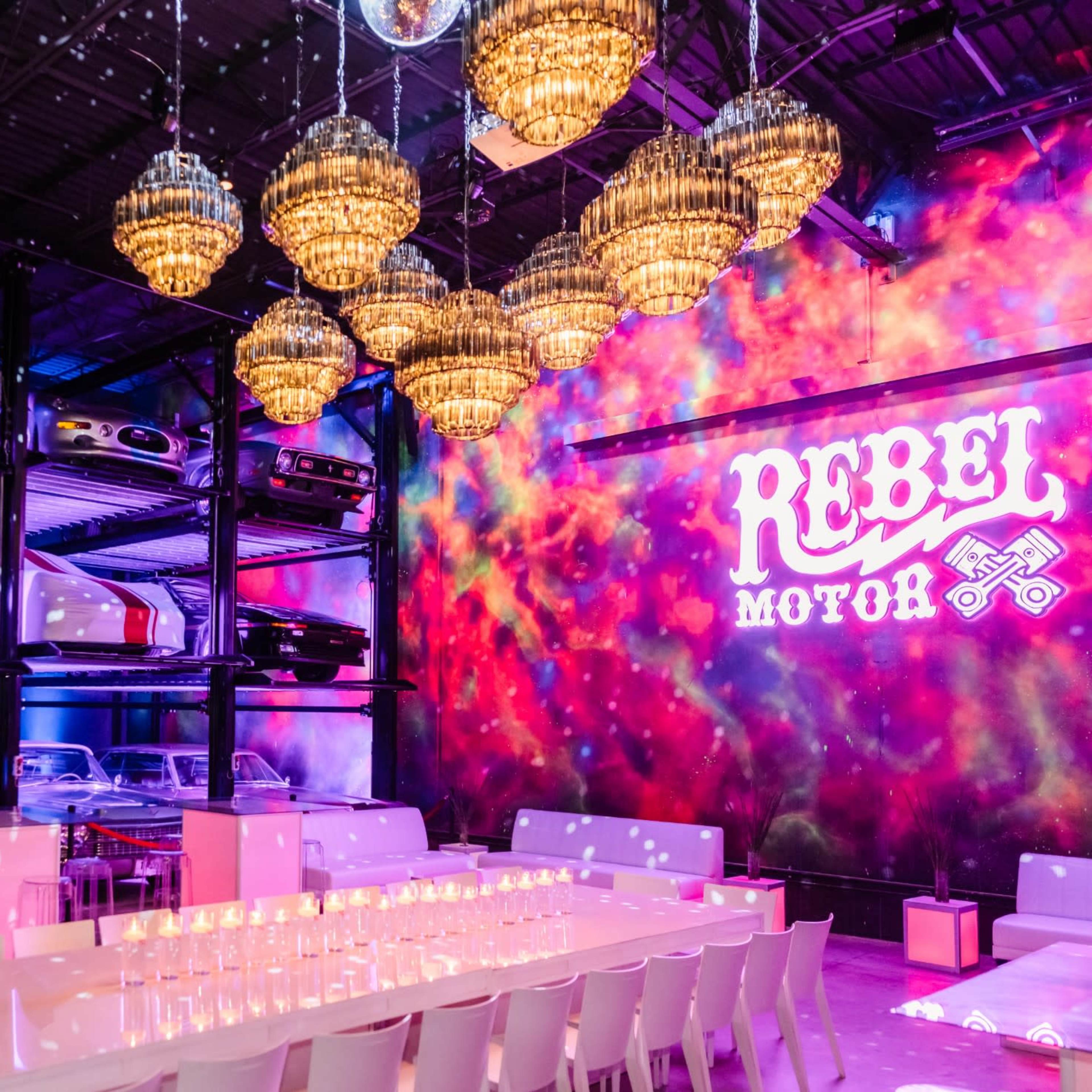 A modern event space featuring a row of luxury cars displayed on shelves, with a long table set for dining beneath decorative chandeliers and a colorful wall mural.