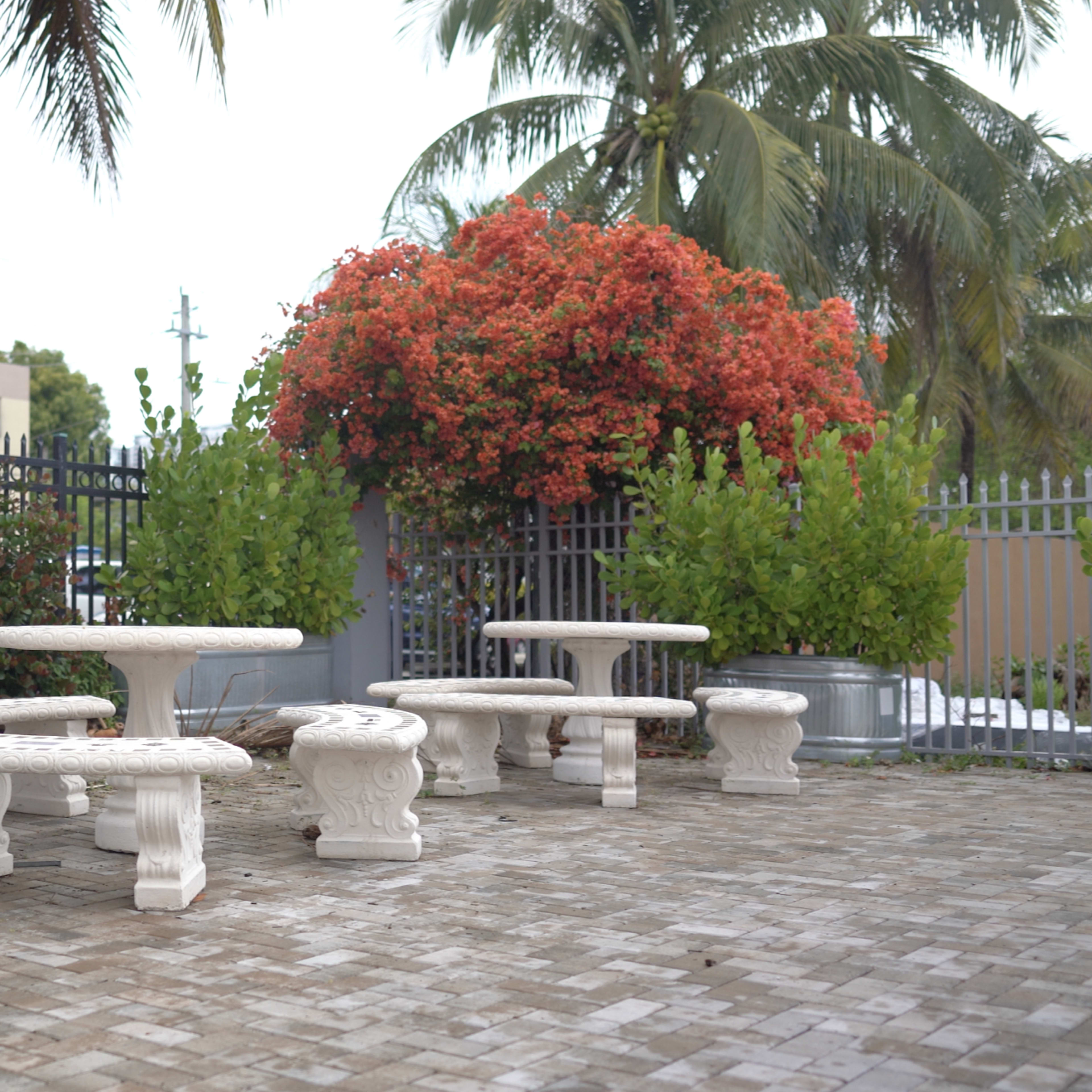 A small outdoor seating area with white stone tables and benches surrounded by greenery and flowering shrubs.