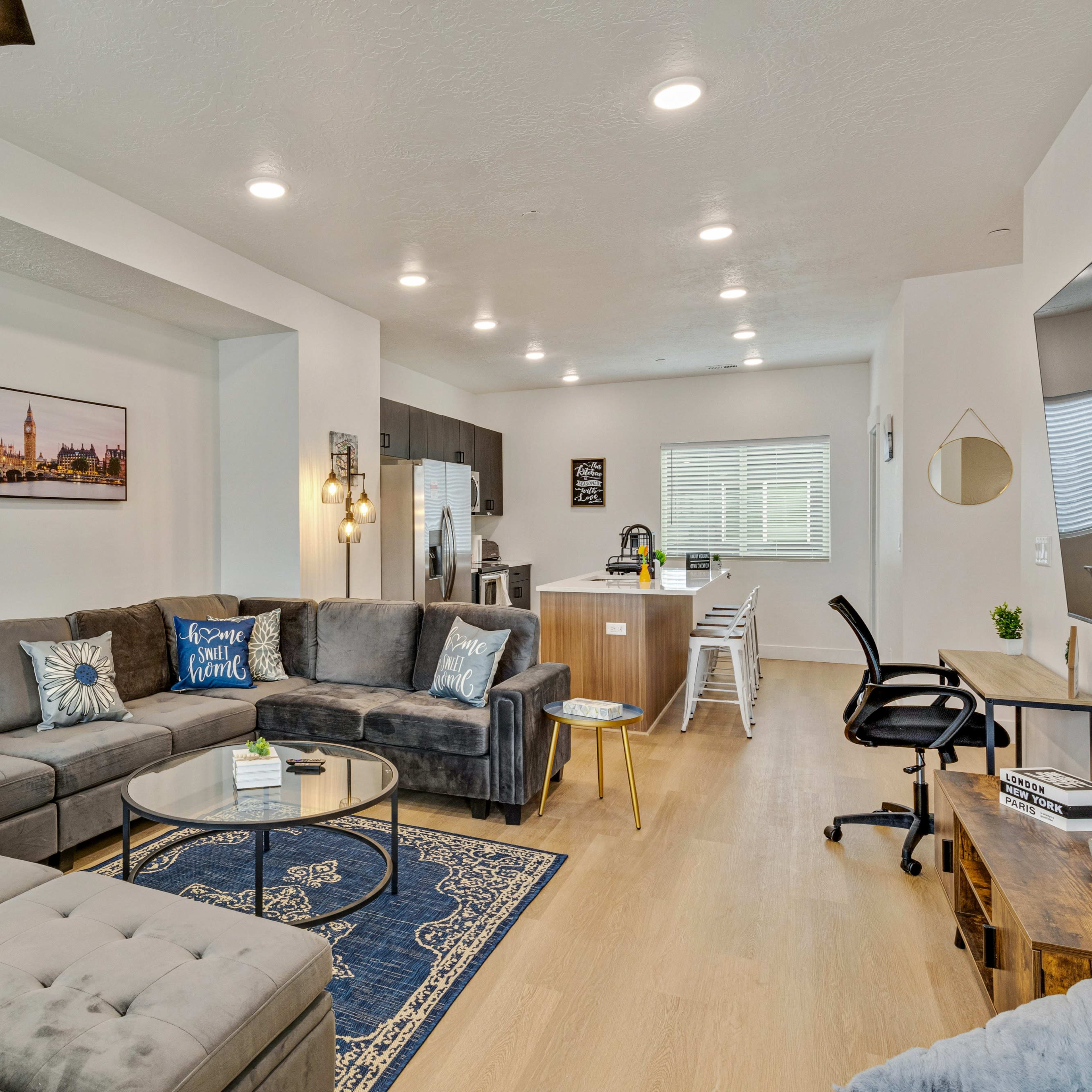 A modern living room with a gray sectional sofa, a round glass coffee table, and a kitchen area in the background.