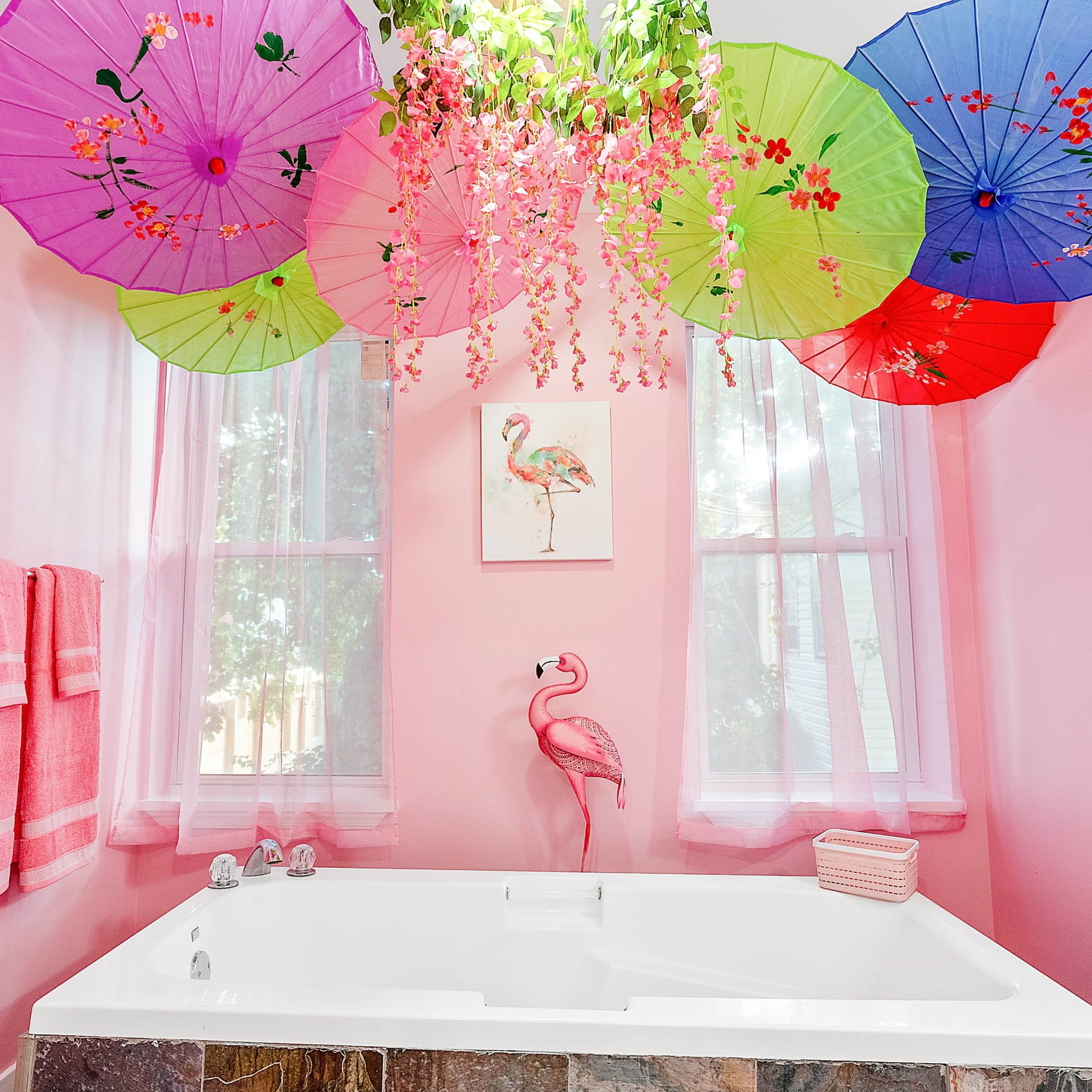 A pink bathroom features a large bathtub under colorful umbrellas and floral decorations, with a pink flamingo display on the wall and pink towels hanging nearby.