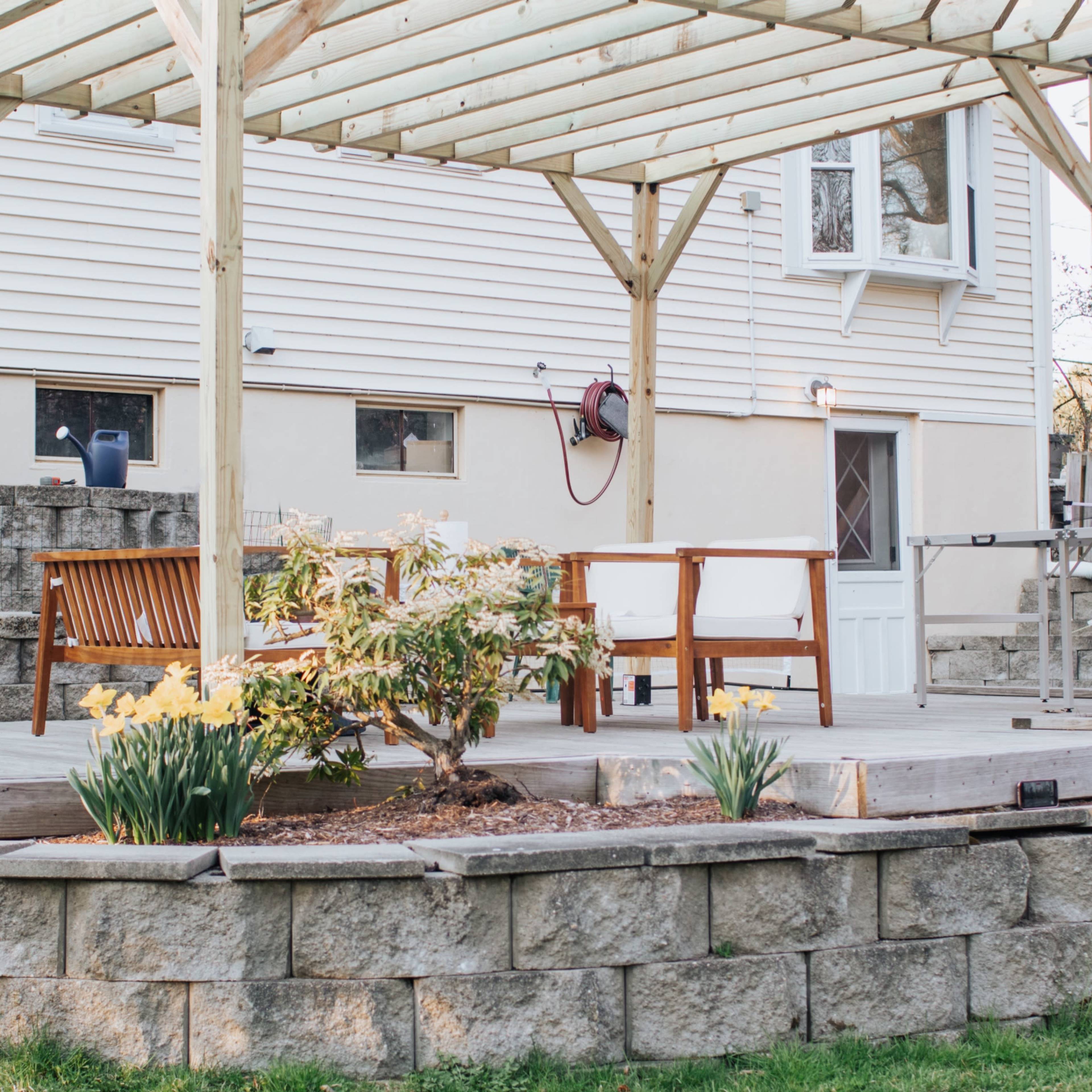 A wooden patio with a pergola features several outdoor furniture pieces and potted plants in a residential backyard.