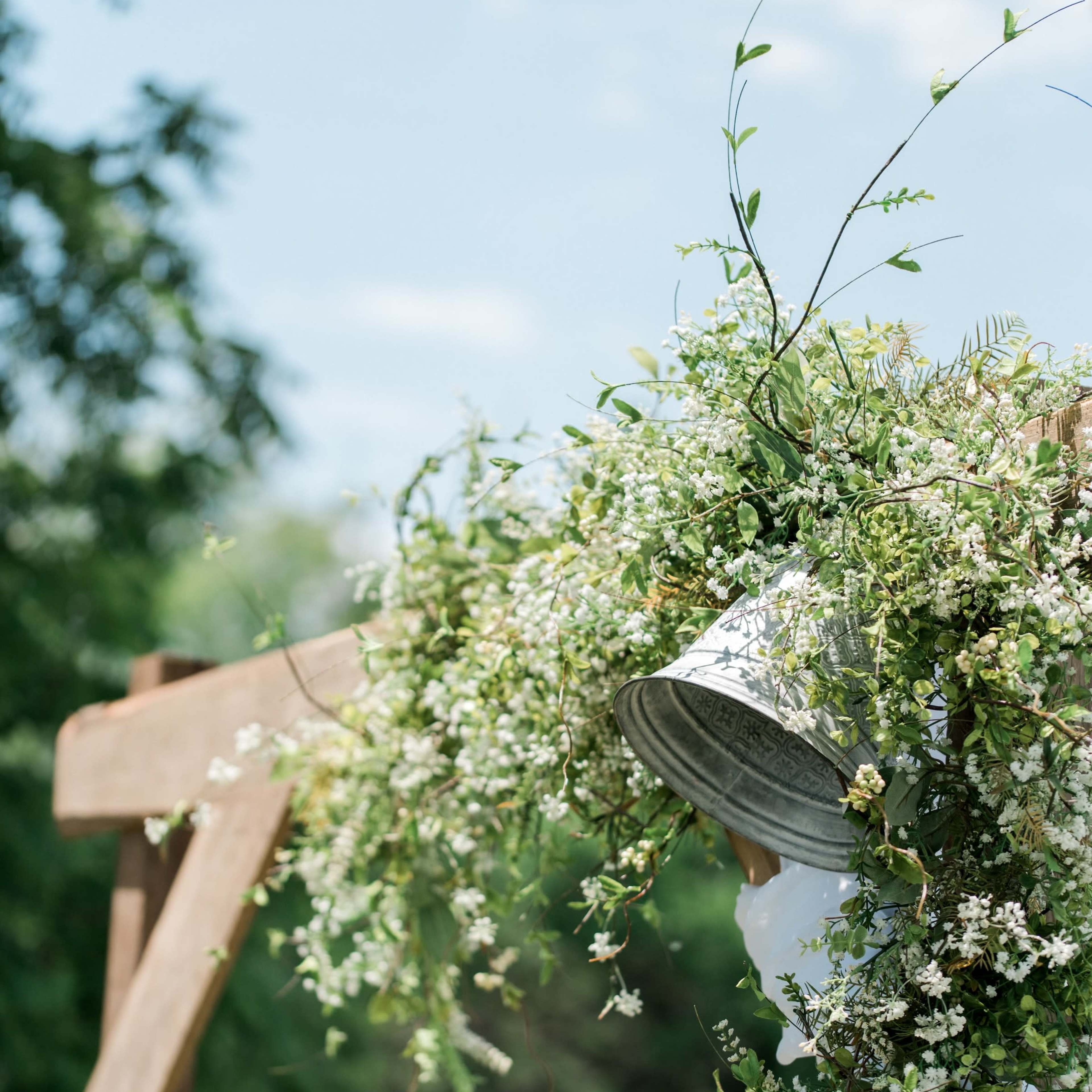 A metal bucket overflows with greenery and small white flowers, hanging from a wooden structure outdoors.
