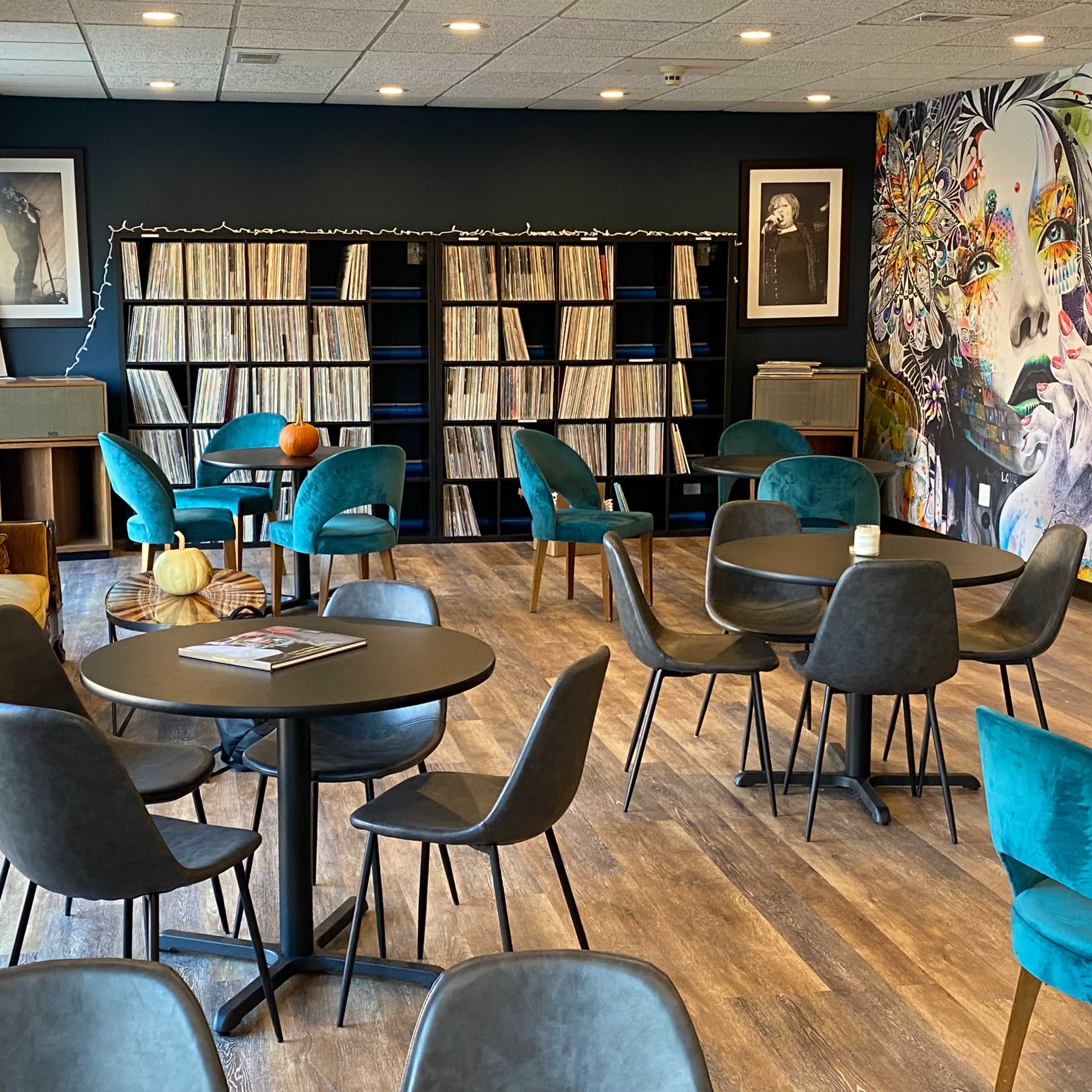 A cozy lounge area featuring tables and chairs, a vinyl record shelf, and a wall adorned with a colorful mural.