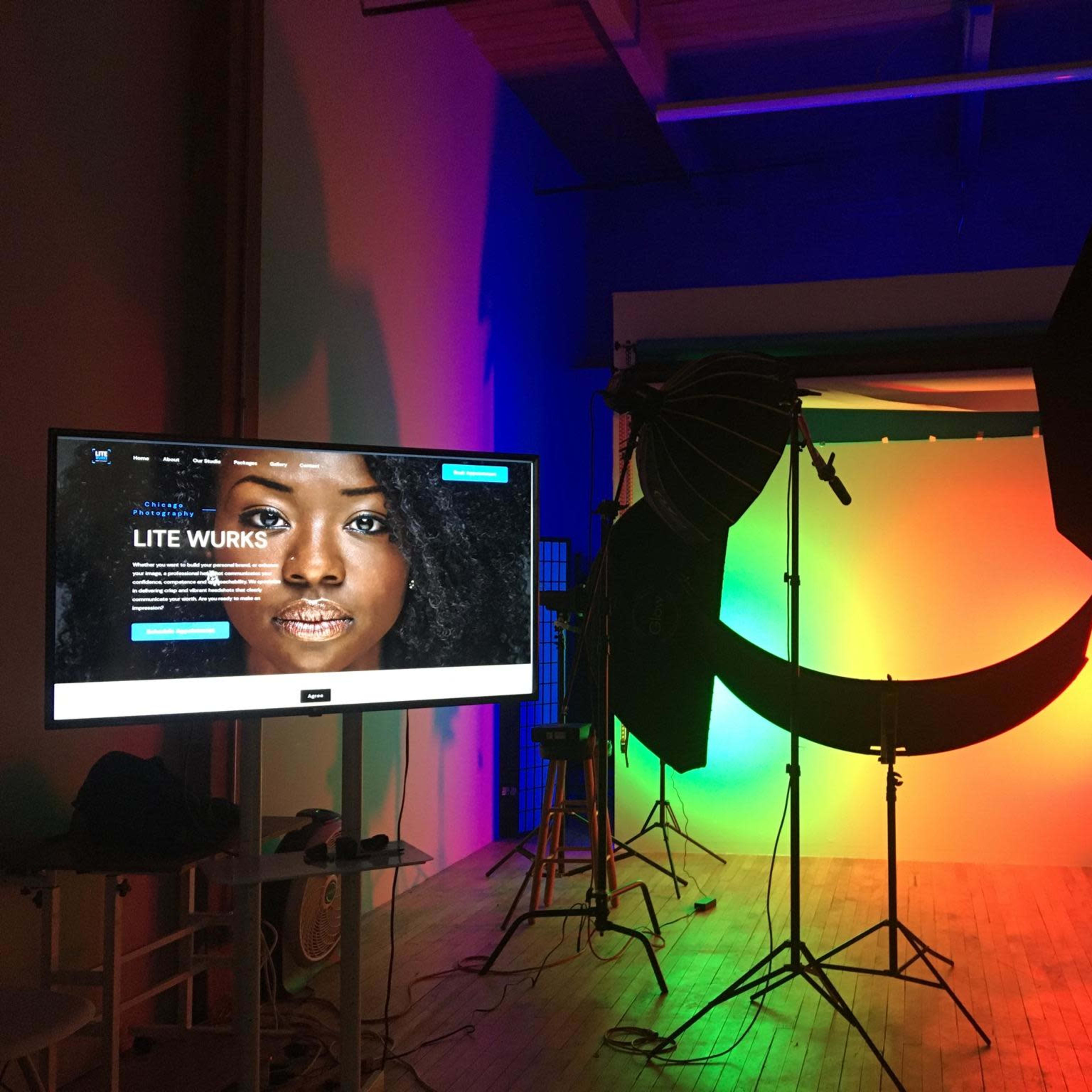 A photography studio setup with colorful lighting, a backdrop, and a monitor displaying a website.