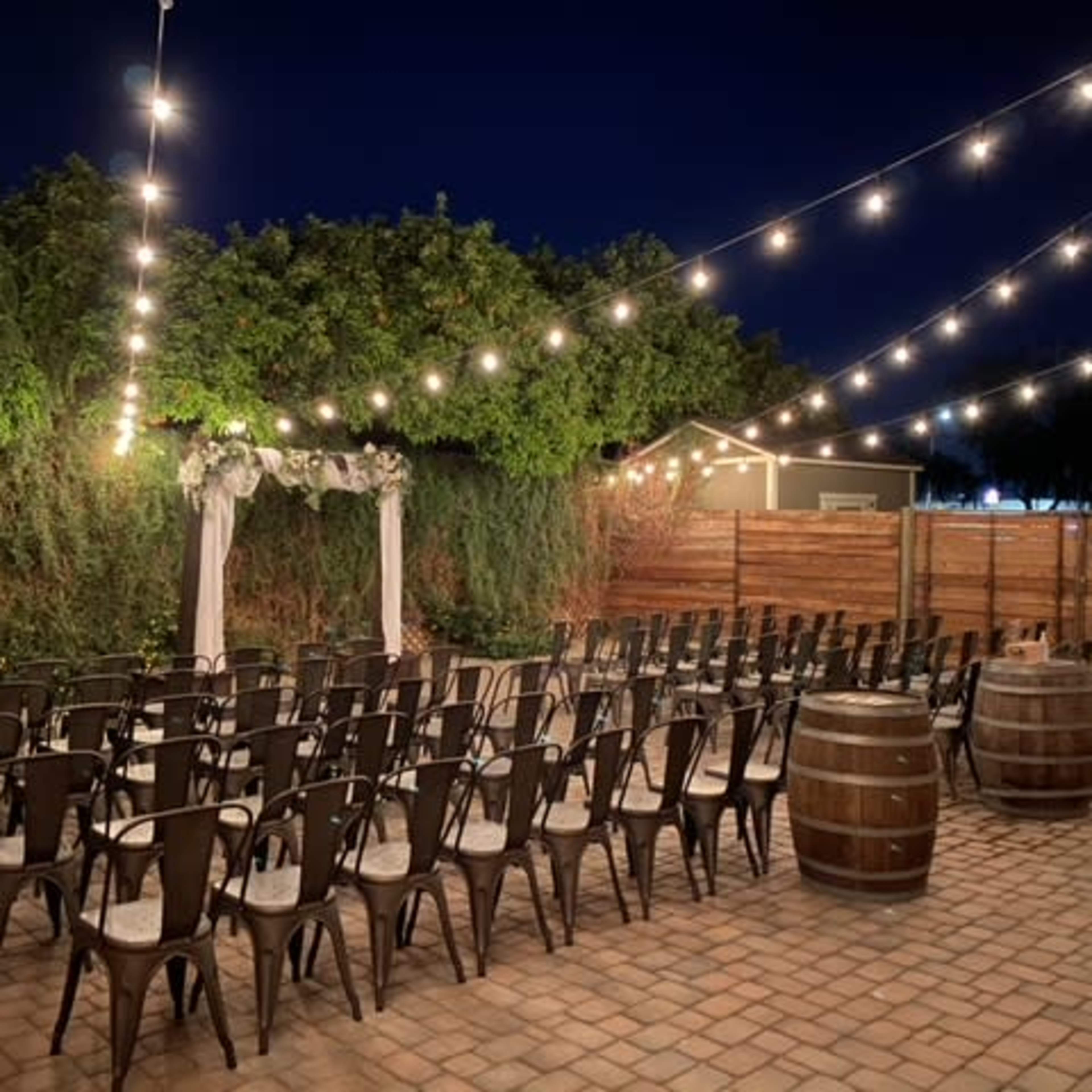An outdoor event space at night, featuring rows of chairs arranged facing an archway, with string lights overhead and wooden barrels nearby.