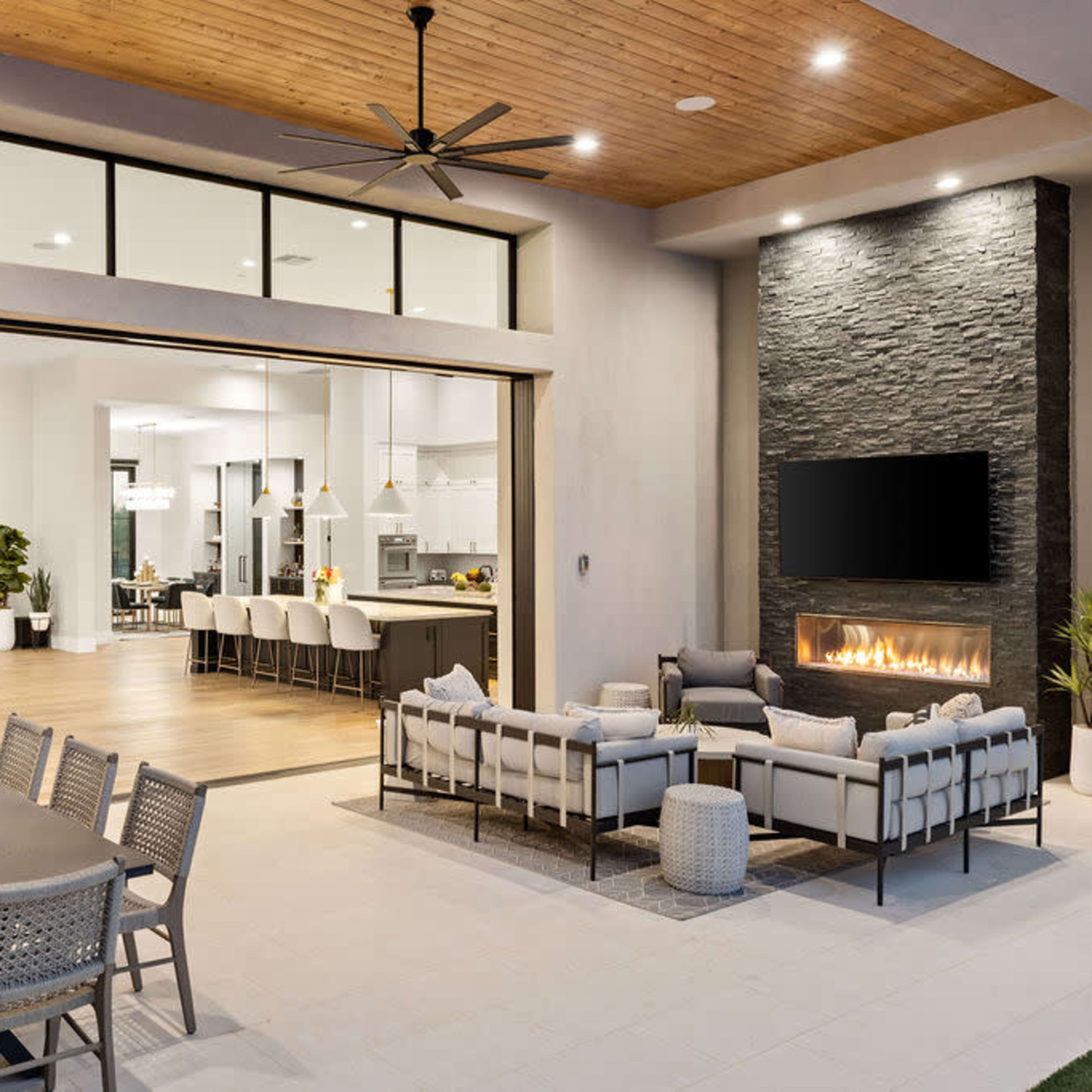 A modern outdoor living space with a seating area and a dining table, adjacent to an open kitchen with large windows and a stone fireplace.