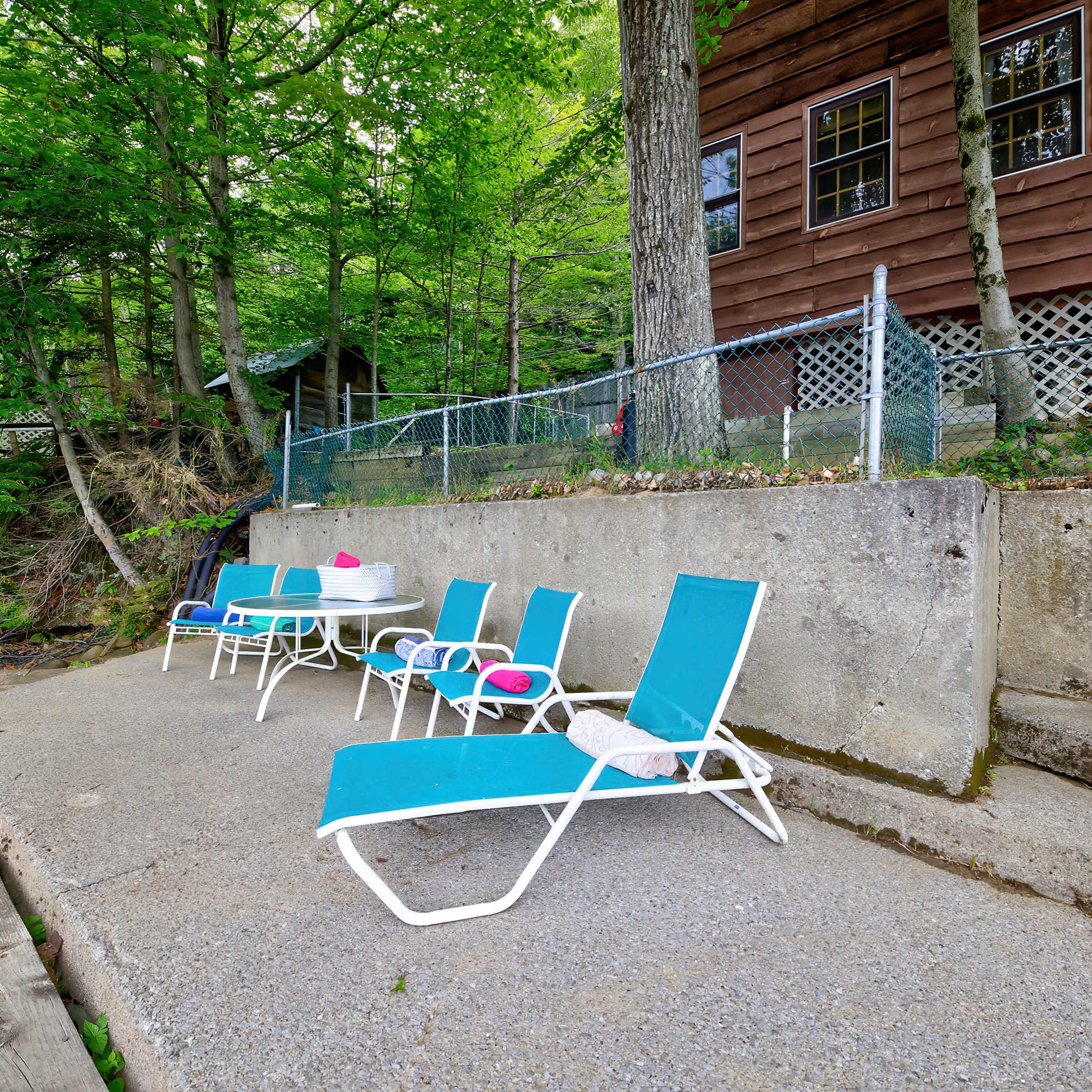 A concrete patio with several blue lounge chairs and a table is situated next to a wooded area and a rustic cabin.