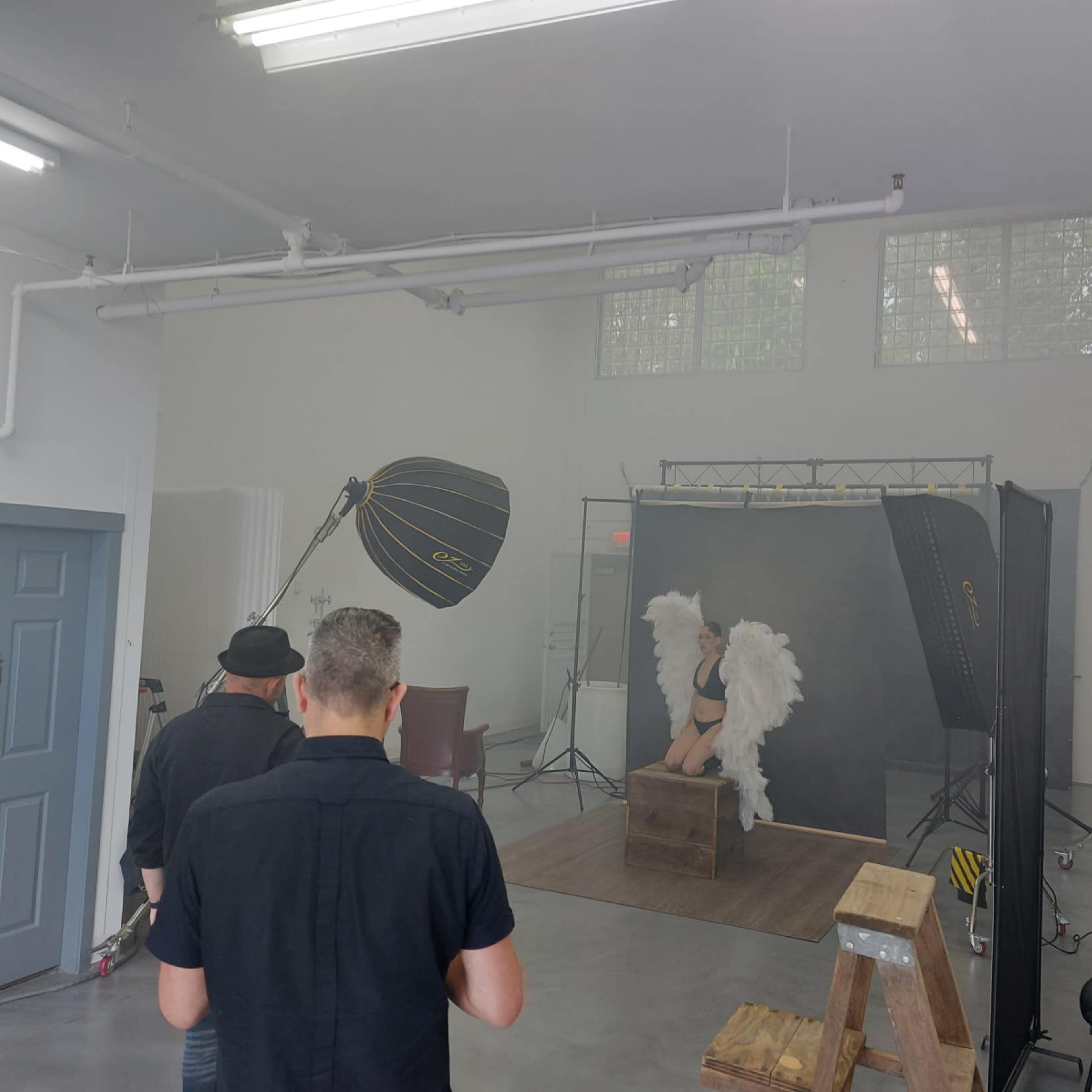A photoshoot setup features a model with wings posing on a wooden box, while photographers adjust equipment in a spacious studio.