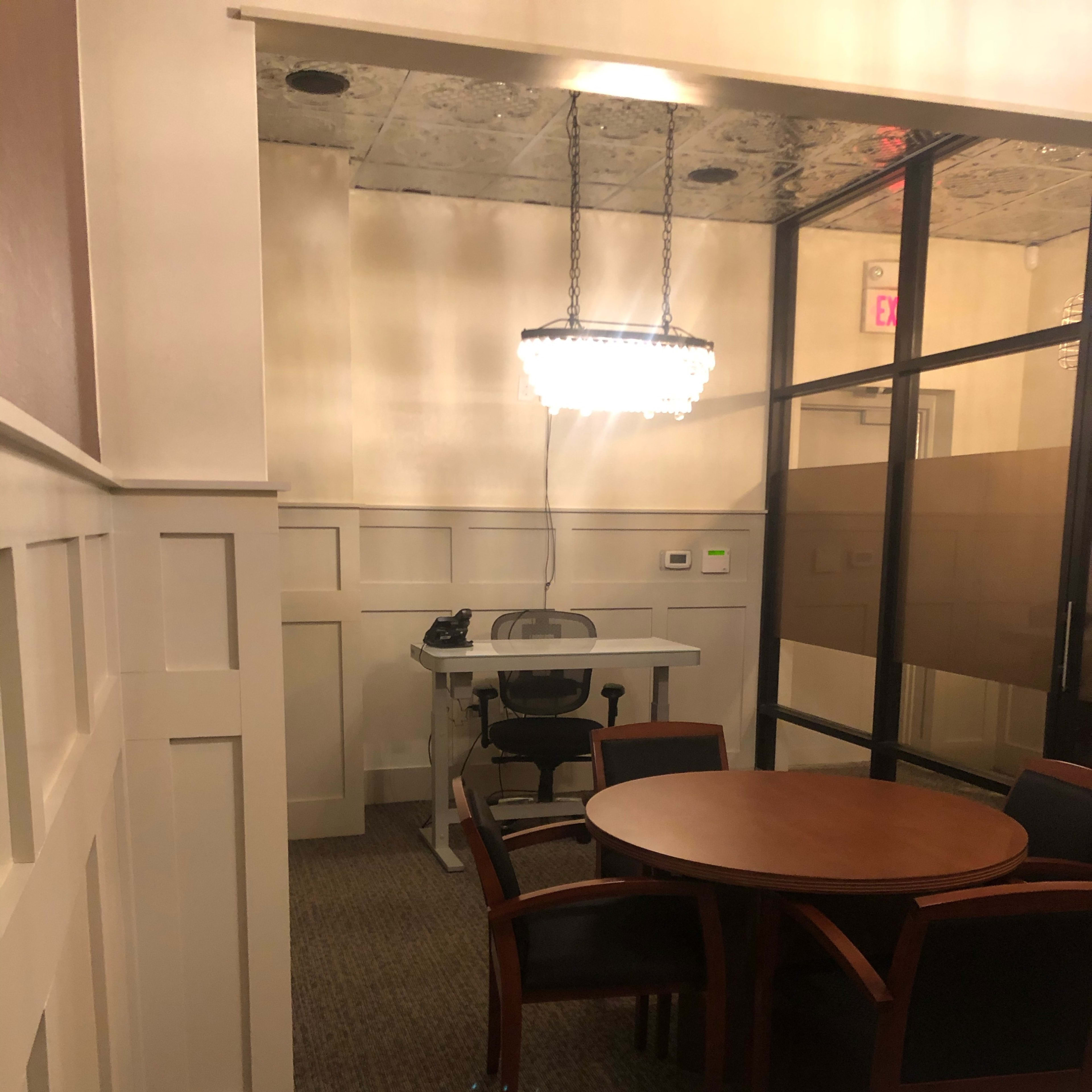 A conference room with a round table and chairs, a desk with an office chair, and a chandelier above.