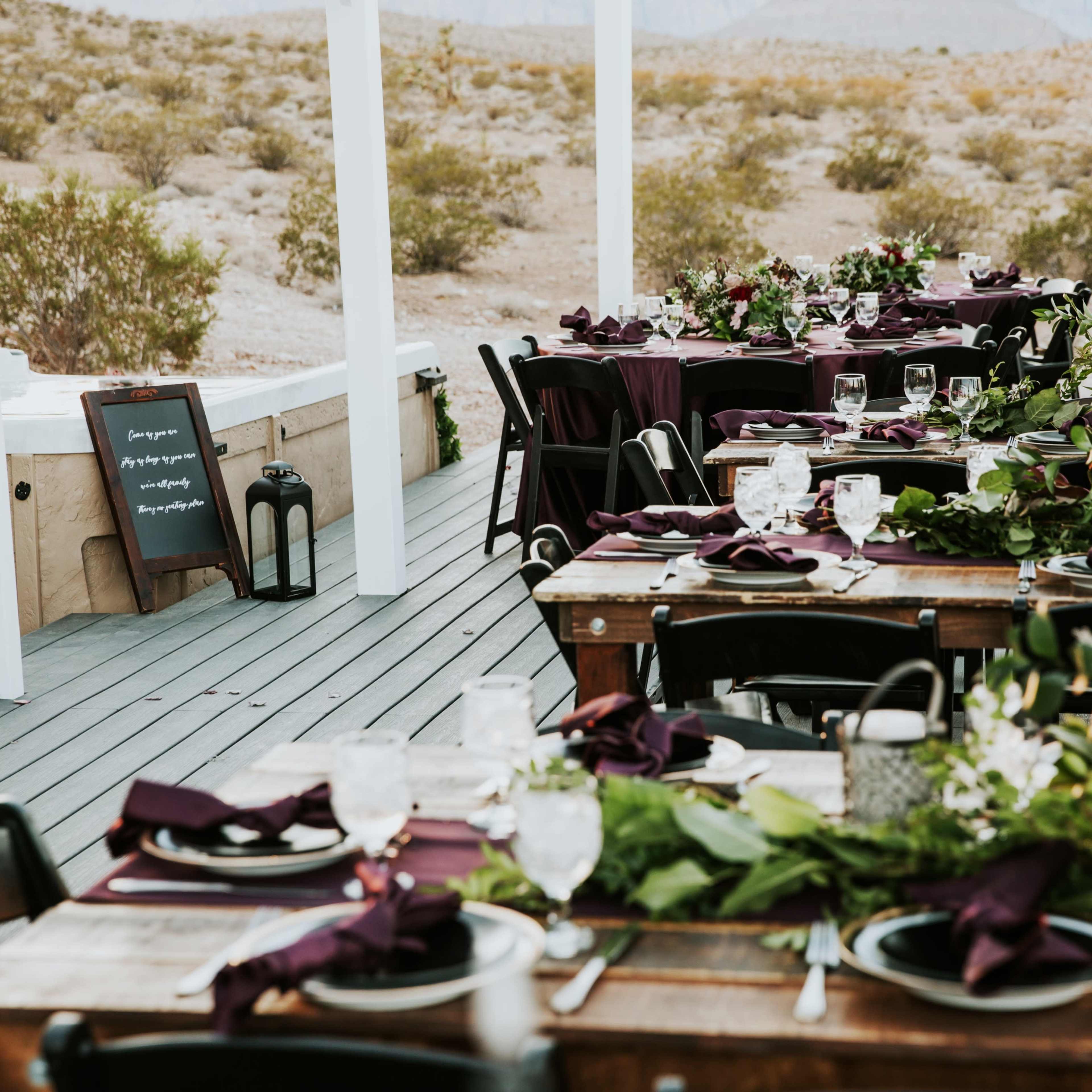 A set of elegantly arranged tables with dark tablecloths and floral centerpieces is placed on a wooden deck overlooking a desert landscape.