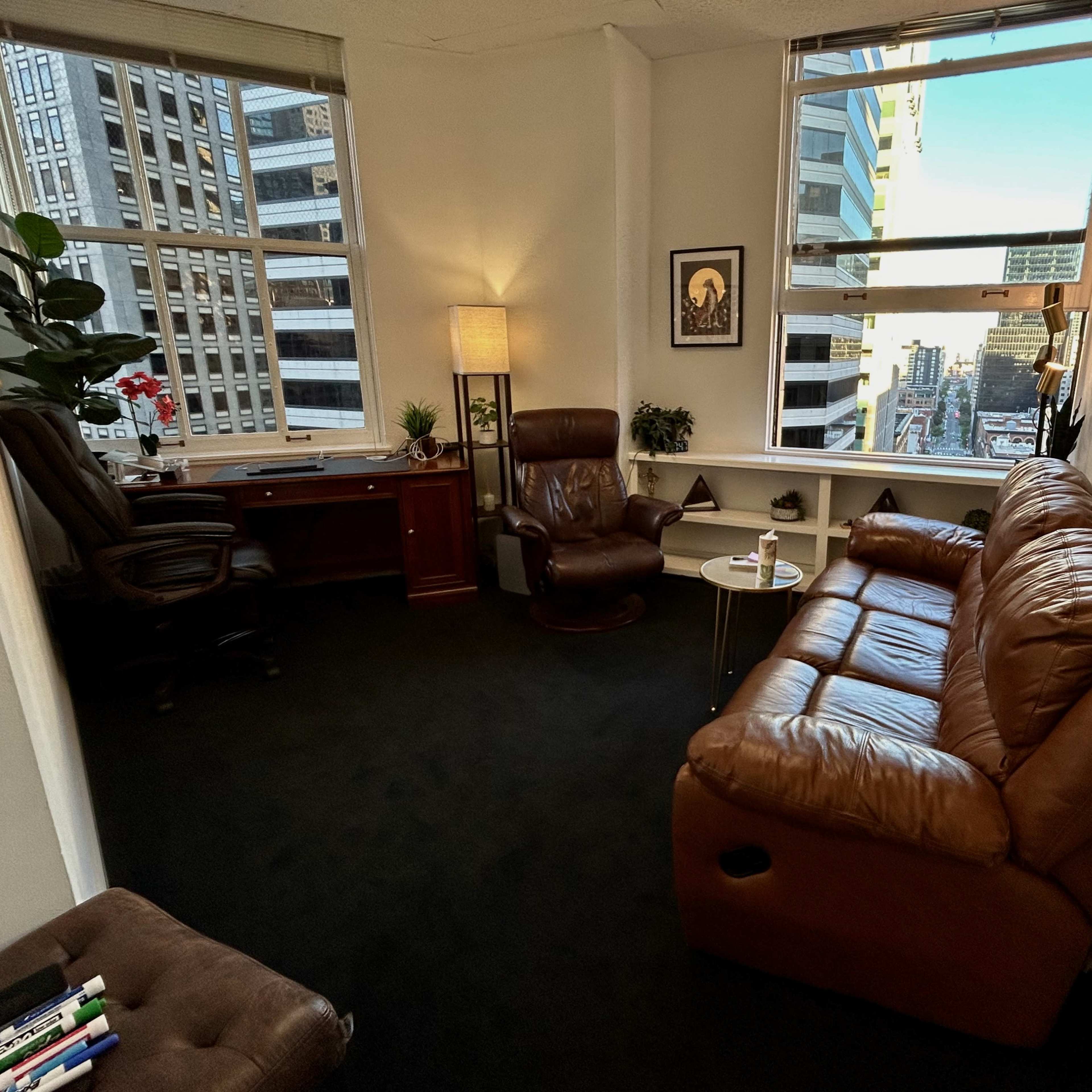 A corner office with a brown leather sofa, an armchair, a desk with a computer, and large windows offering a view of a cityscape.