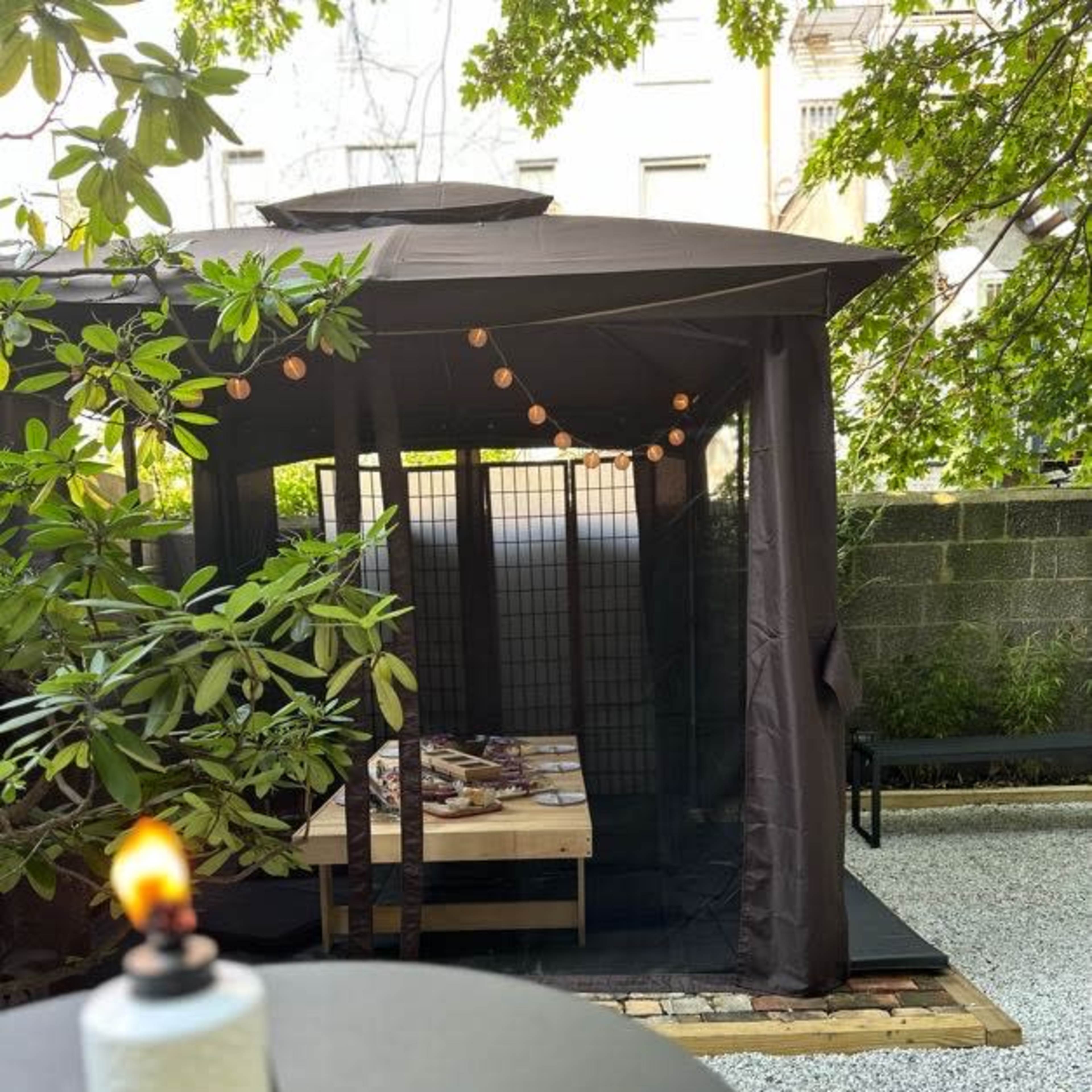 A dimly lit outdoor gazebo with string lights and a wooden platform is set within a landscaped area featuring gravel and greenery.