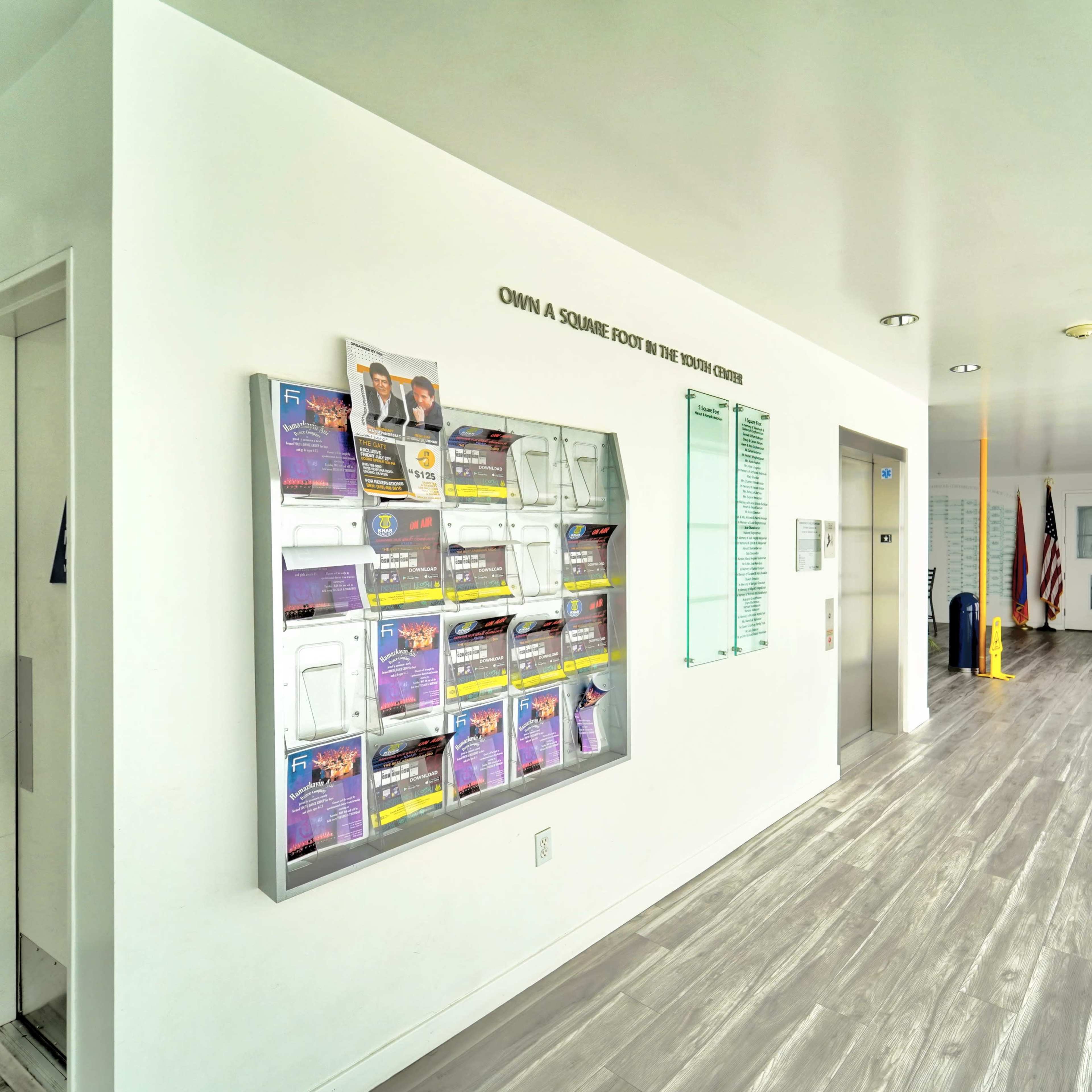 A bright, modern hallway with a bulletin board displaying flyers on the left, an elevator door, and a clear view of the entrance at the far end.