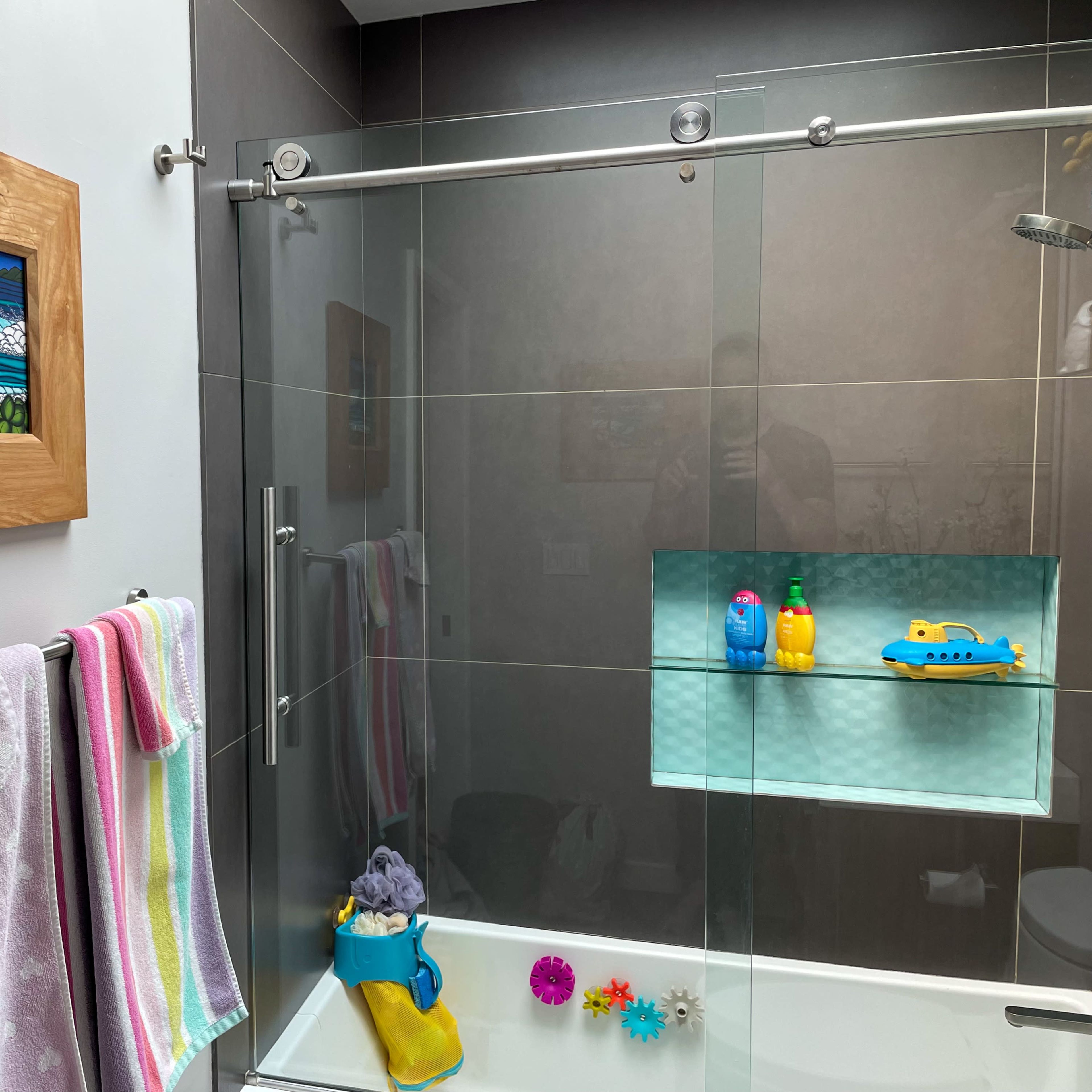A modern bathroom featuring a glass shower enclosure, a wooden-framed artwork on the wall, and colorful bath toys arranged on a shelf.