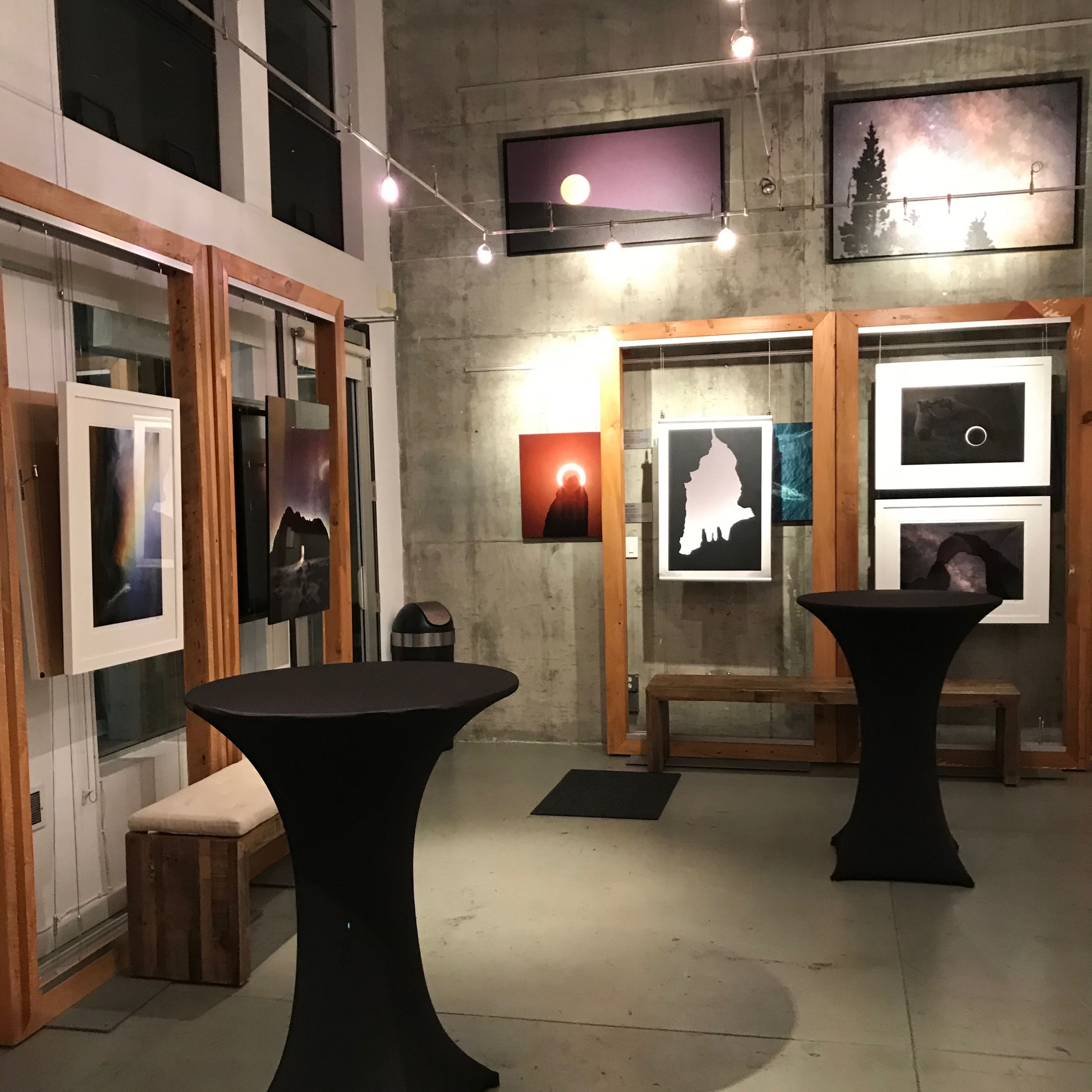 An art gallery featuring framed photographs displayed on walls, with two tall black tables in the center under soft overhead lighting.
