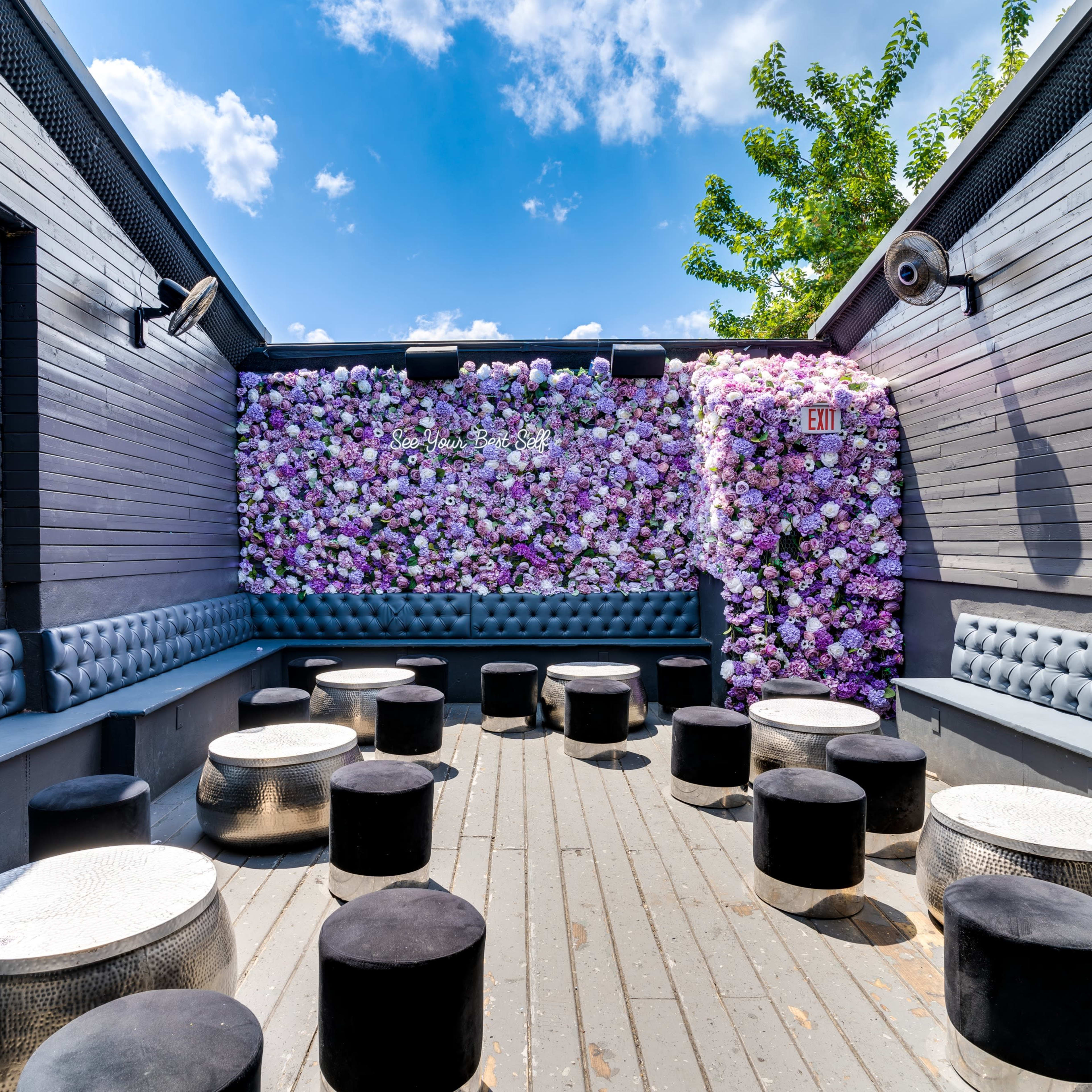 A stylish outdoor seating area features a wall of flowers and circular stools against a backdrop of blue sky and greenery.