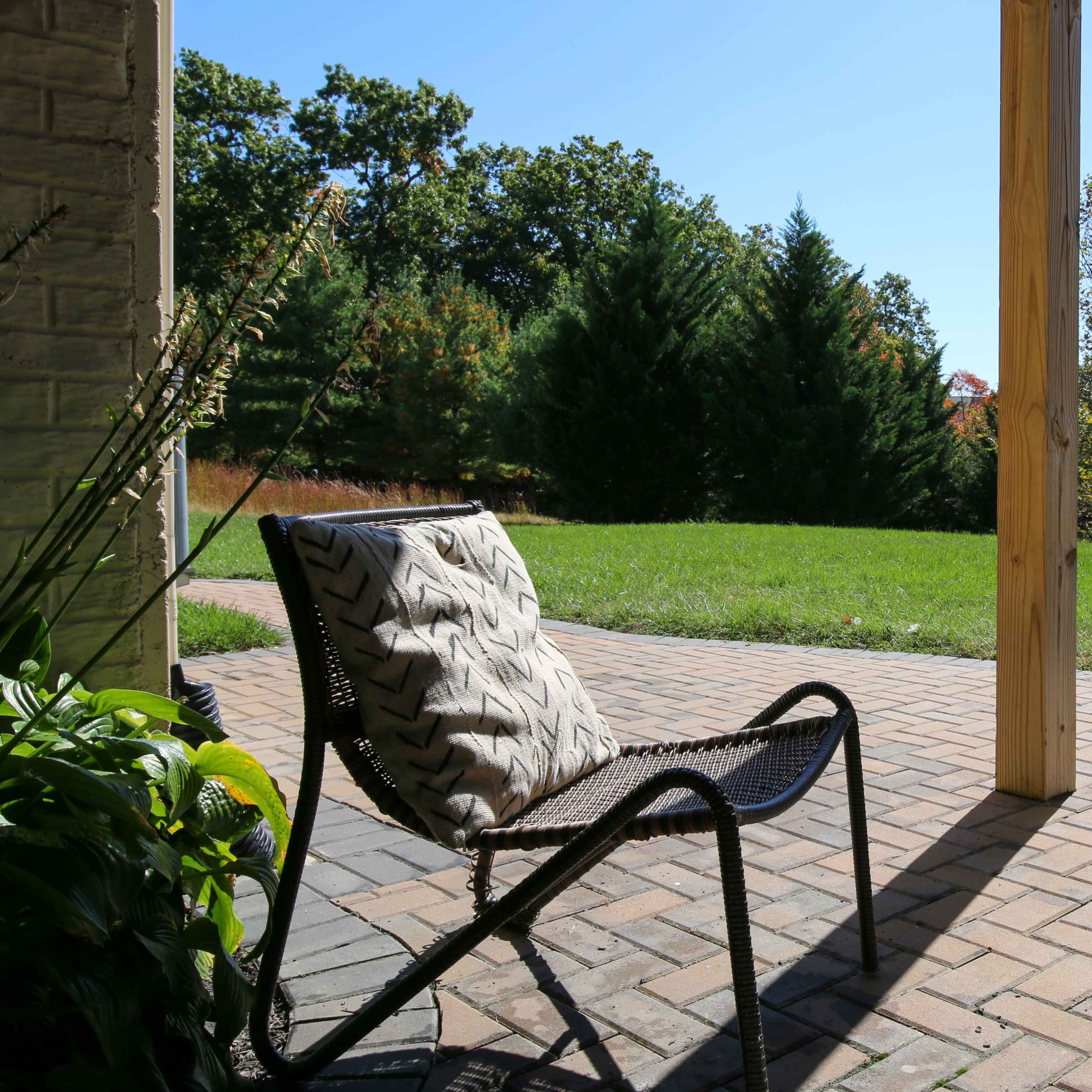 A chair with a patterned cushion is positioned on a brick patio, overlooking a grassy area and trees in the background under a clear sky.