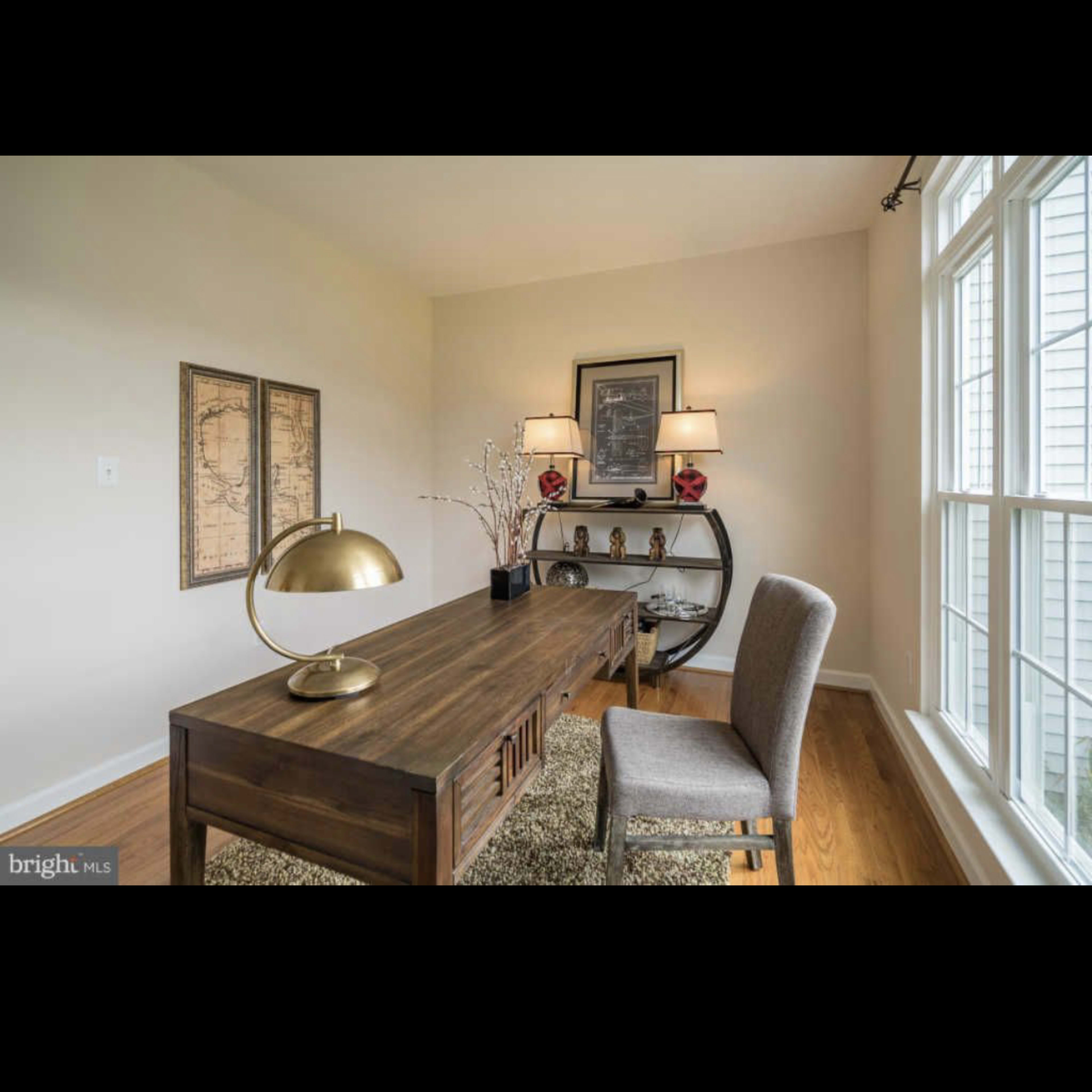 A modern home office features a wooden desk with a lamp, a chair, and decor items against a backdrop of large windows.