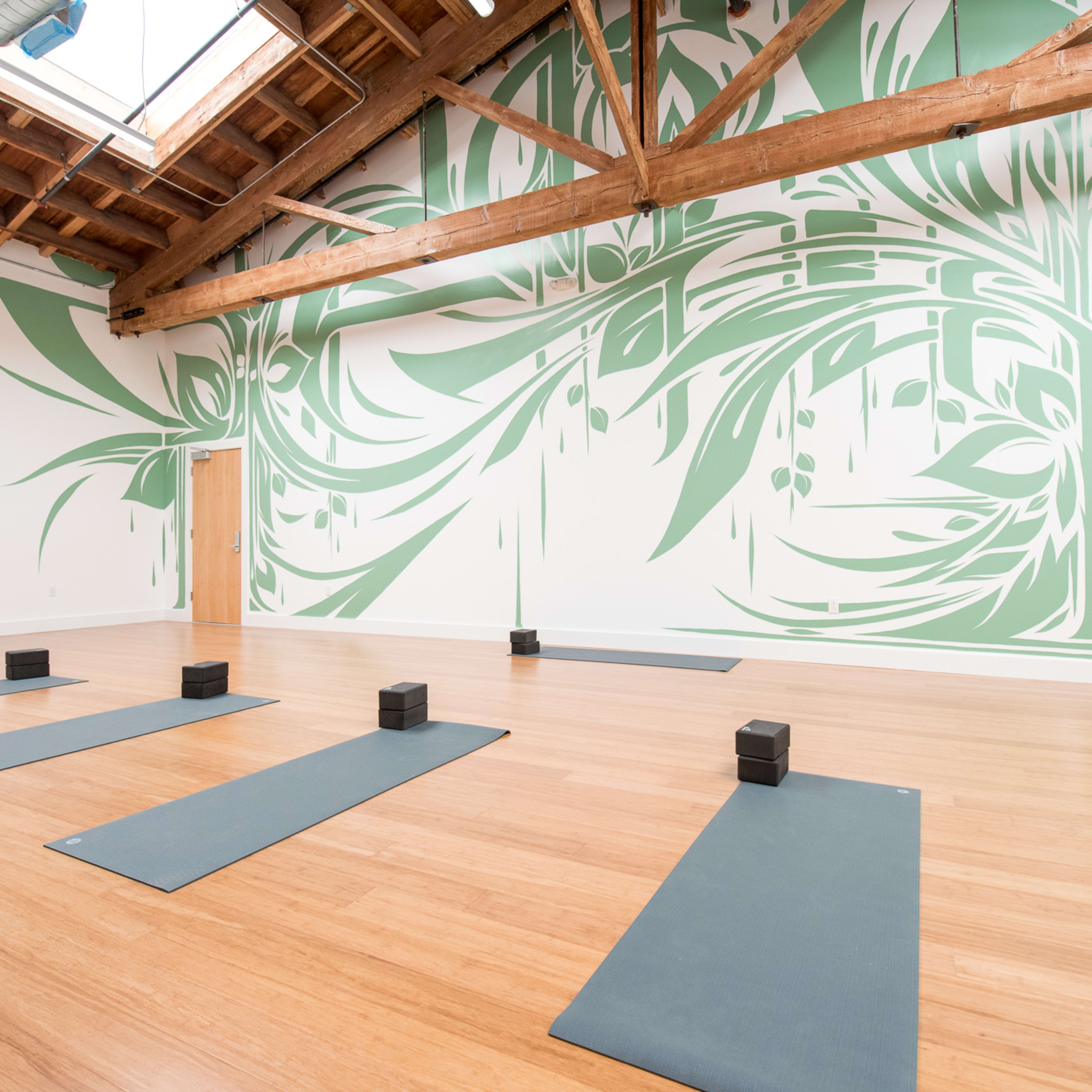 A spacious yoga studio with bamboo flooring, six yoga mats arranged on the floor, and a green mural on one wall.