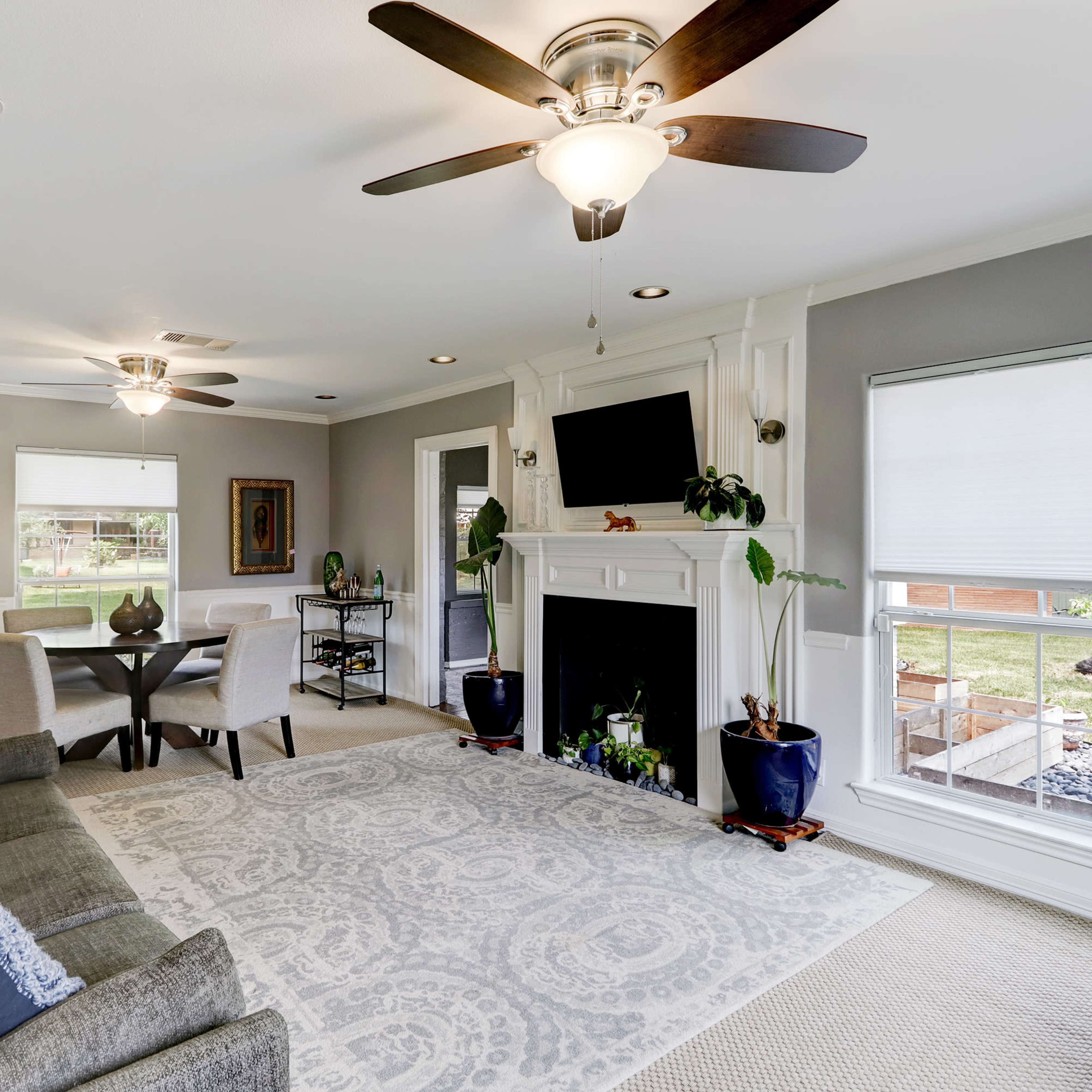 A living room with a gray and white color scheme, featuring a seating area, a fireplace, and a view of a backyard through large windows.