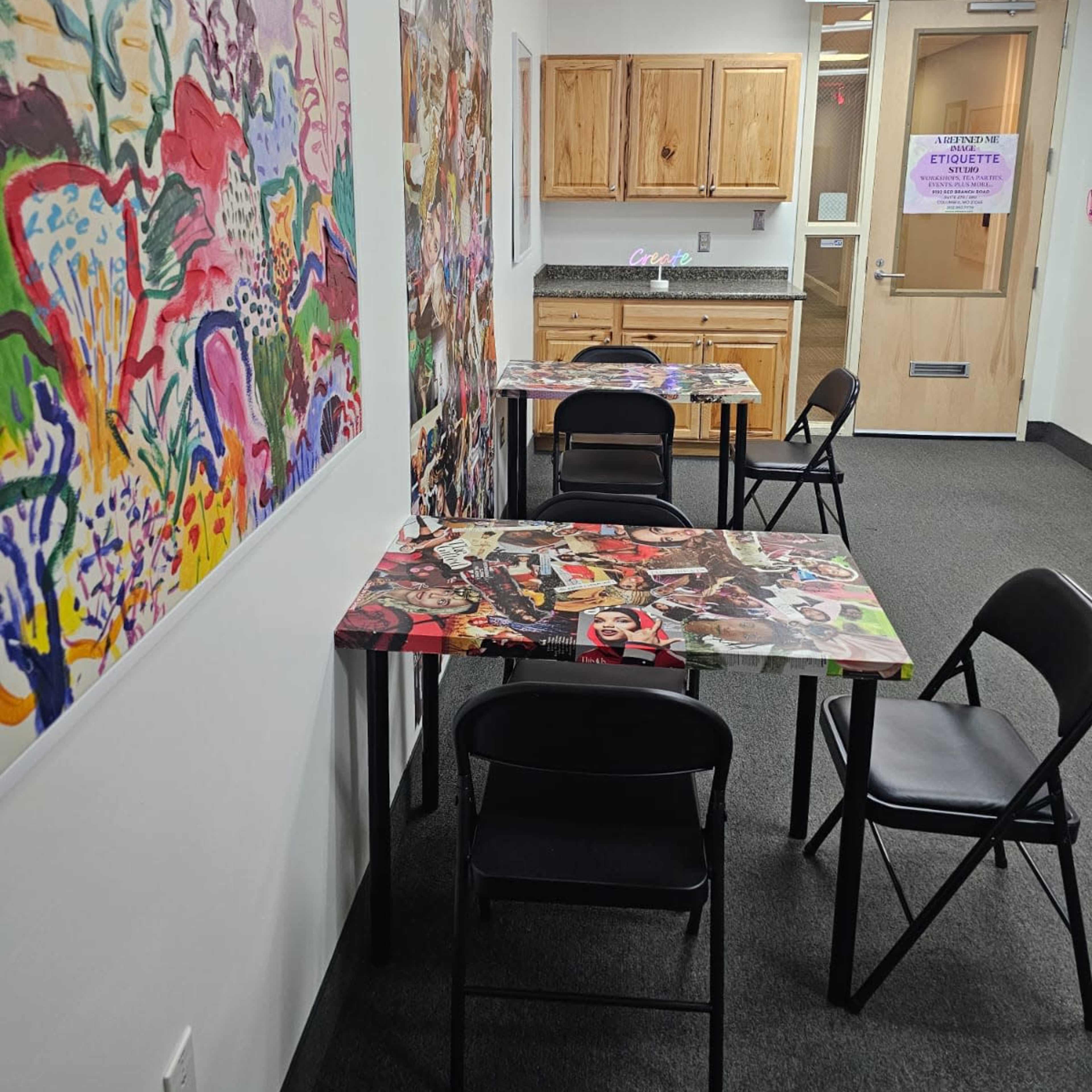 A narrow hallway with four tables covered in artwork and chairs, alongside colorful wall art and a kitchenette area in the background.