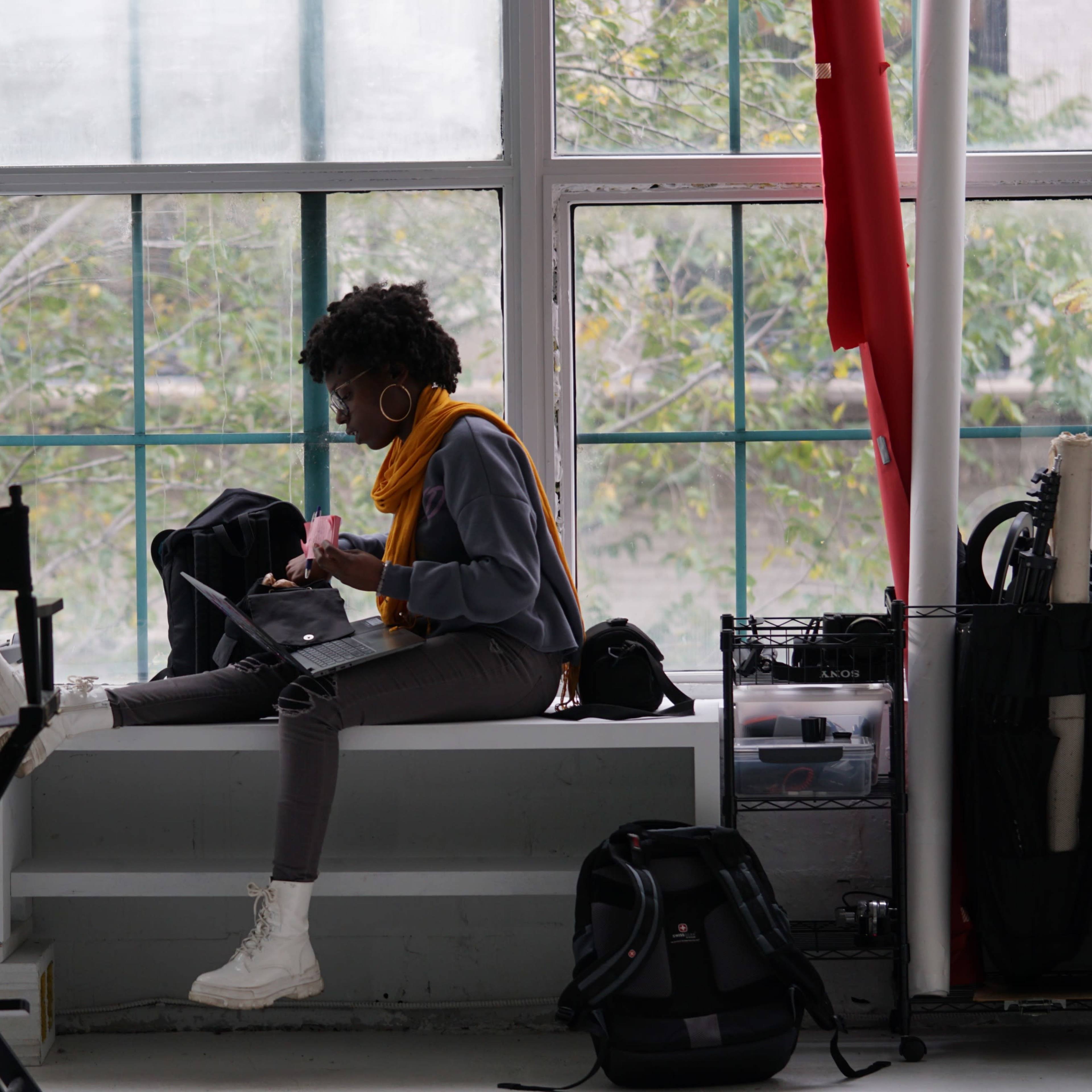 A person sits on a bench by a large window, working on a laptop surrounded by bags and equipment.
