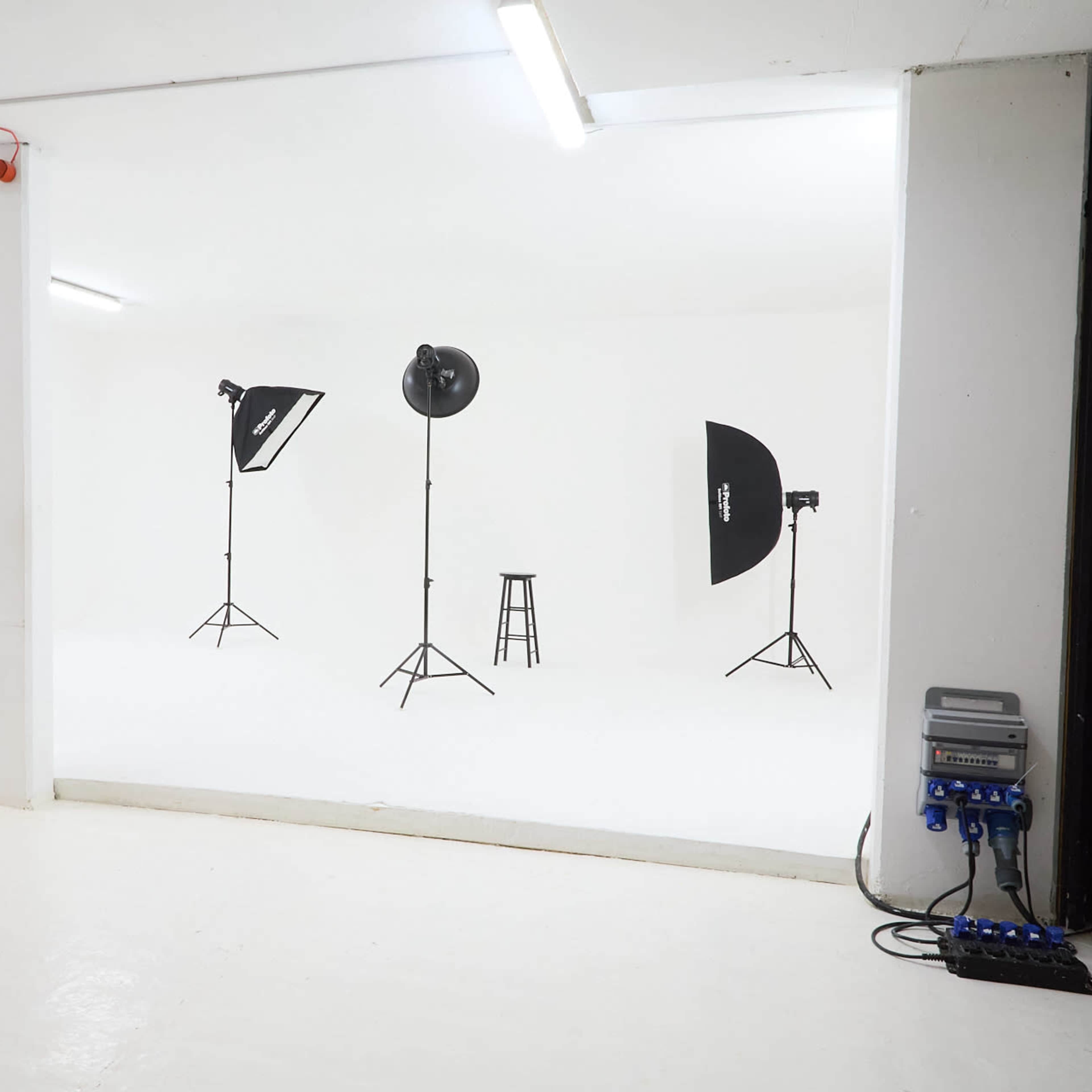 A photography studio with a white backdrop, three light stands, and a stool positioned on a white floor.